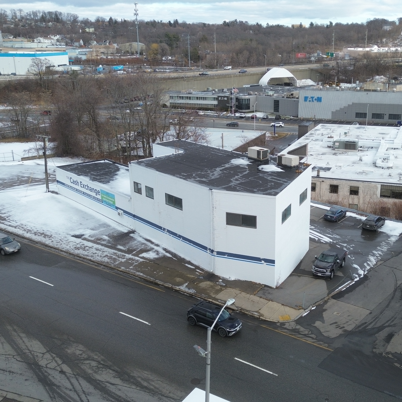 A white building with a sign indicating it is 'The Cash Exchange' situated at a corner in an urban area. The building has a flat roof with patches of snow, a small parking lot with vehicles, and a street with a few cars in front. Snow is on the groun
