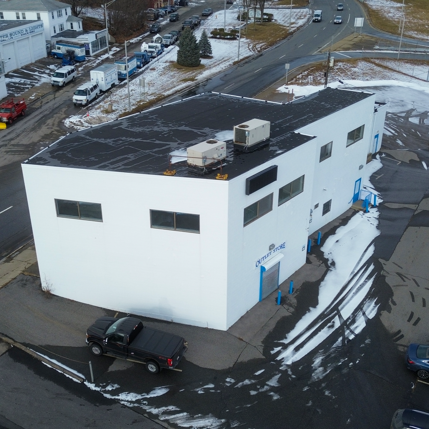 An aerial view of a white two-story building marked 'Outlet Store' with a black roof, surrounded by parking lots with some snow, multiple parked vehicles, and a few trees, in a commercial area.