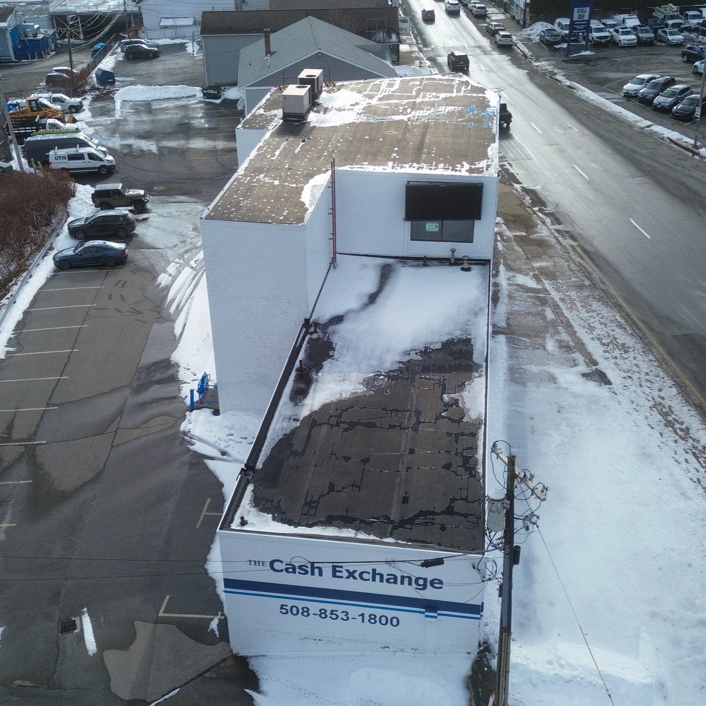 An aerial view of a building with a sign that reads 'The Cash Exchange' and phone number 508-853-1800. The roof is partially covered in snow, with some areas cleared or melting. Surrounding the building are parking lots, snow piles, and a street with
