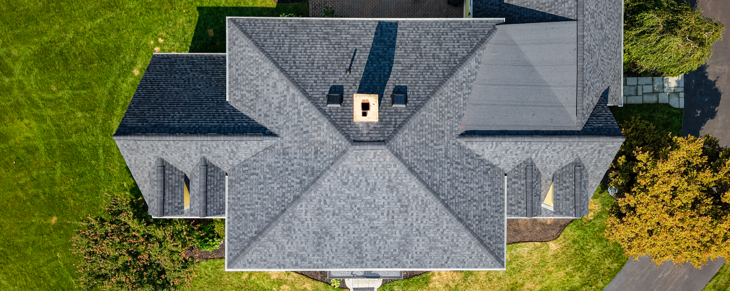 Aerial view of a gray-shingled house with a complex roof design, surrounded by green lawn and trees.