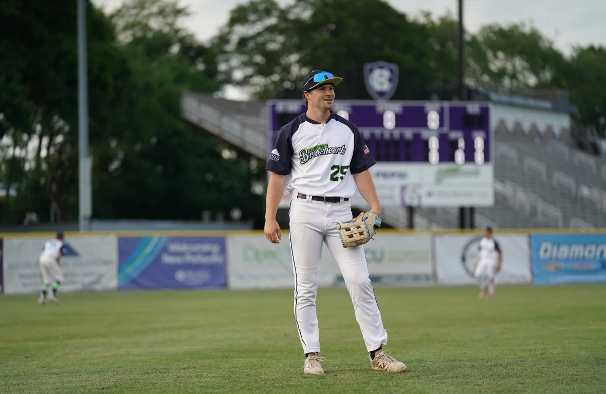 A baseball player standing on the field, wearing a white and black uniform with the number 25, holding a glove, with others in the background and a scoreboard behind.