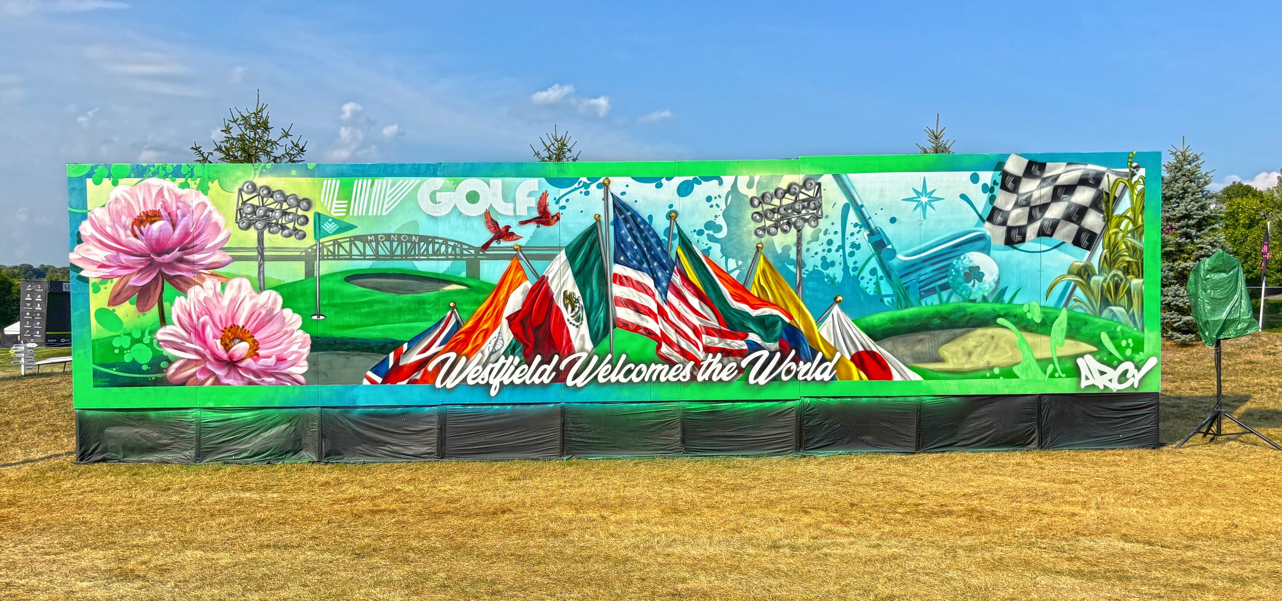 Colorful mural featuring pink flowers, flags from various countries, a golf course, and a checkered flag, with the text 'Westfield Welcomes the World'. ARCY LIV GOLF LIVE EVENT MURAL INDIANAPOLIS INDIANA