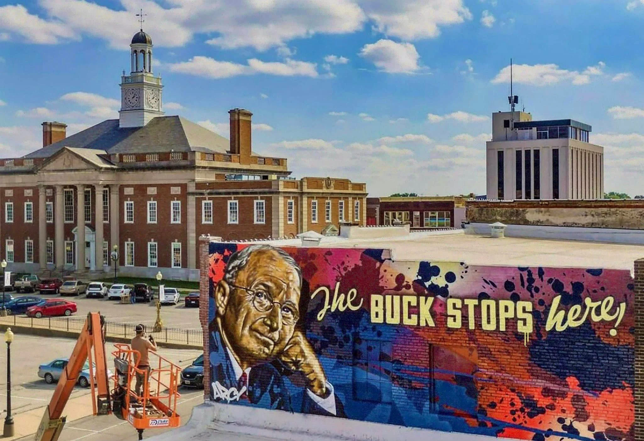 A colorful mural of a man with glasses and gray hair, with the text 'The BUCK STOPS here!' painted on the wall. In the background, there are various buildings, including a brick courthouse with a clock tower and a modern office building, with cars parked in front of the buildings and a man operating a lift next to the mural.
