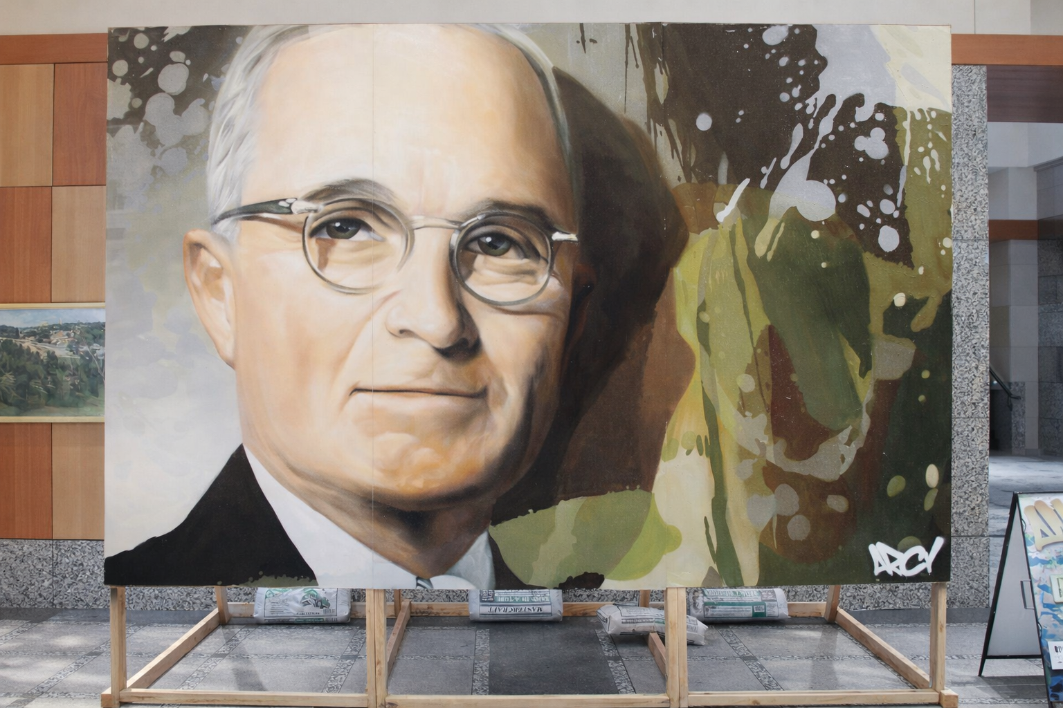 A large portrait painting of an older man wearing glasses and a suit, with a serious expression, mounted on a wooden stand inside a building. HARRY S TRUMAN ARCY LIVE EVENT MURAL NATIONAL ARCHIVES PARTNERSHIP