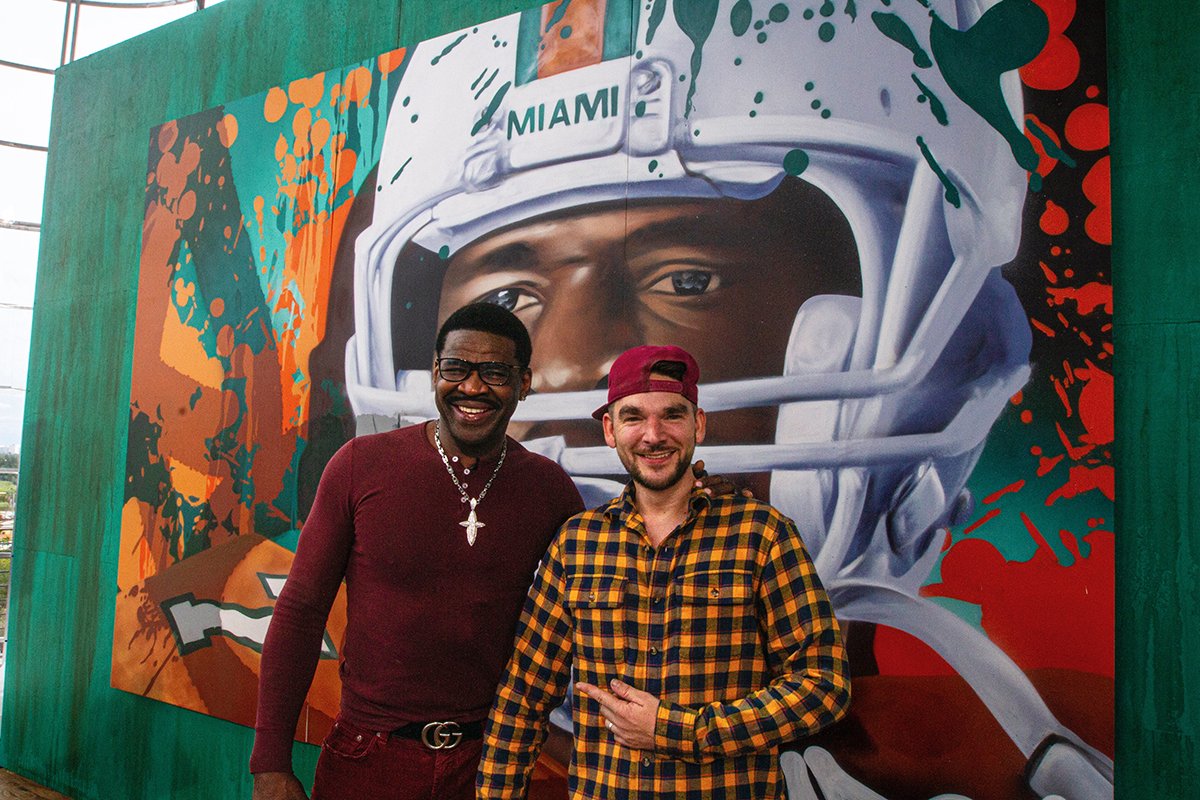 Two men standing in front of a large mural of a football player wearing a helmet with 'MIAMI' written on it. The mural features abstract colors and splash effects. ARCY MICHAEL IRVIN DALLAS COWBOYS UNIVERSITY OF MIAMI THE DAN PATRICK SHOW FLORIDA