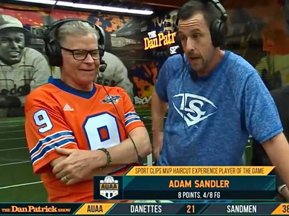 Two men wearing headphones engaged in conversation; one wears an orange sports jersey with the number 9, and the other a blue shirt with a logo; sports-themed backdrop with a banner reading "The Dan Patrick Show". ADAM SANDLER ARCY MURALS