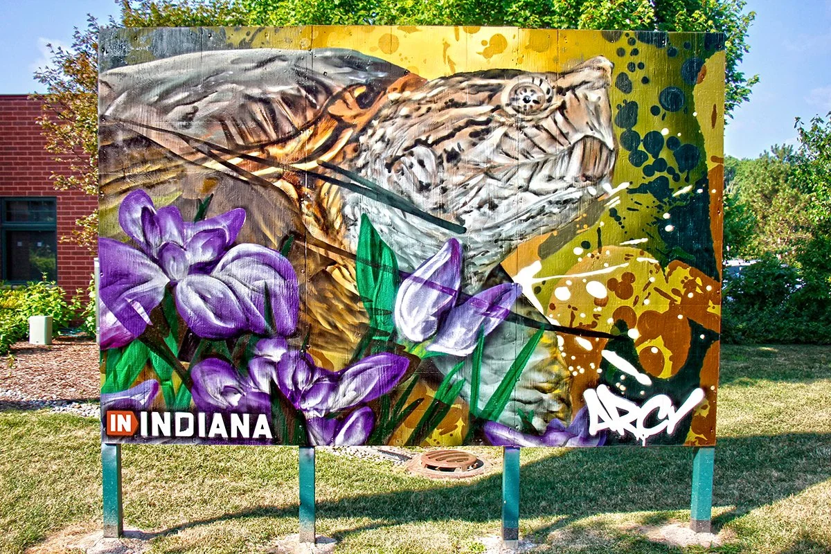 A colorful mural of a tiger with purple flowers and green leaves in the foreground, and the words 'In Indiana' at the bottom. ARCY LIVE EVENT MURAL PORTER INDIANA DUNES