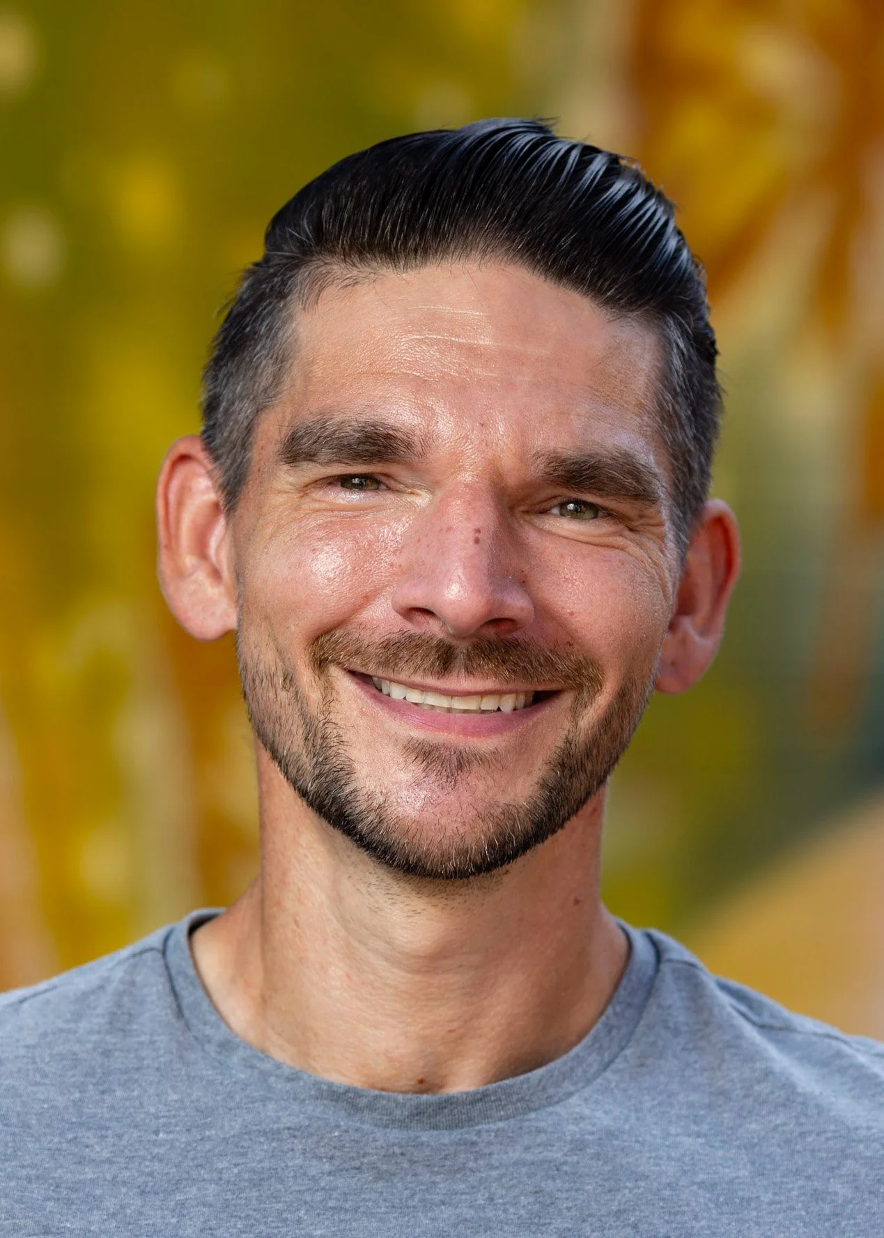 Close-up of a smiling man with dark slicked-back hair, light beard, wearing a gray t-shirt, outdoors with blurred yellow and orange leaves in the background.