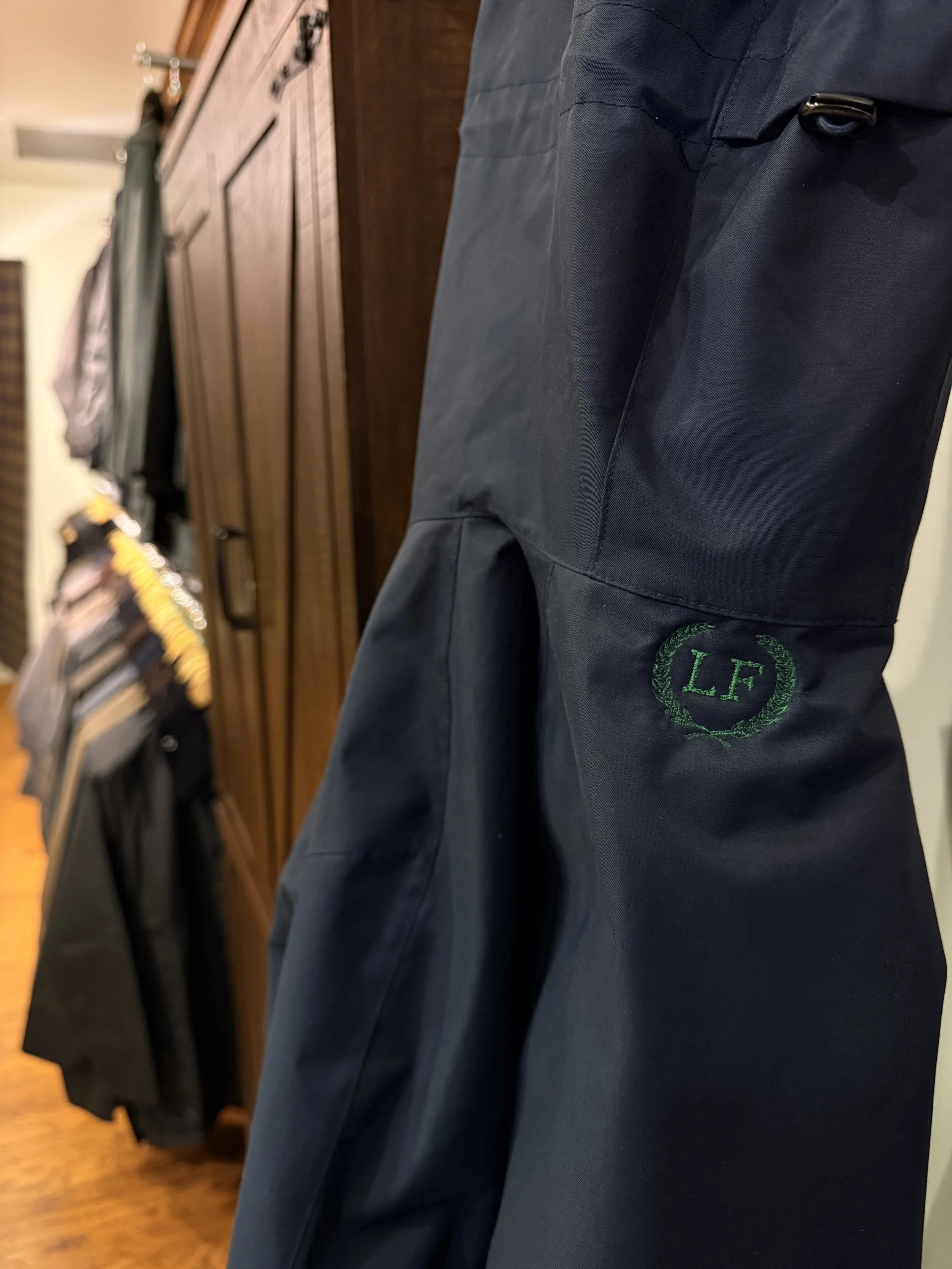 Close-up of dark navy blue pants with green embroidered initials "LF" inside a laurel wreath, hanging on a rack in a clothing store with other clothing in the background.