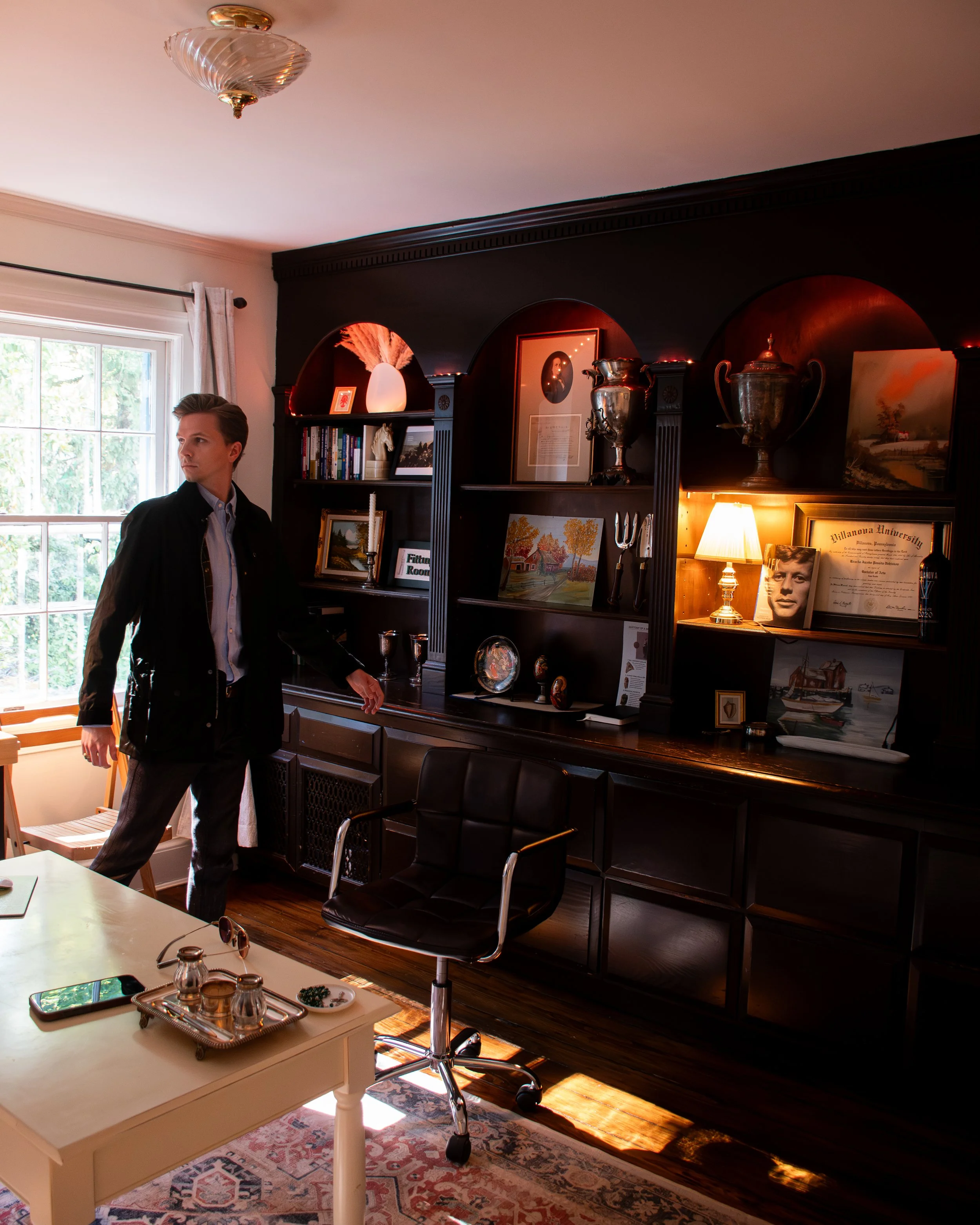 A young man in a black jacket and light blue shirt standing in a room with a dark wooden bookshelf, framed art, trophies, and a lamp. There is a window with light-colored curtains, and the room features hardwood floors and an ornate area rug.