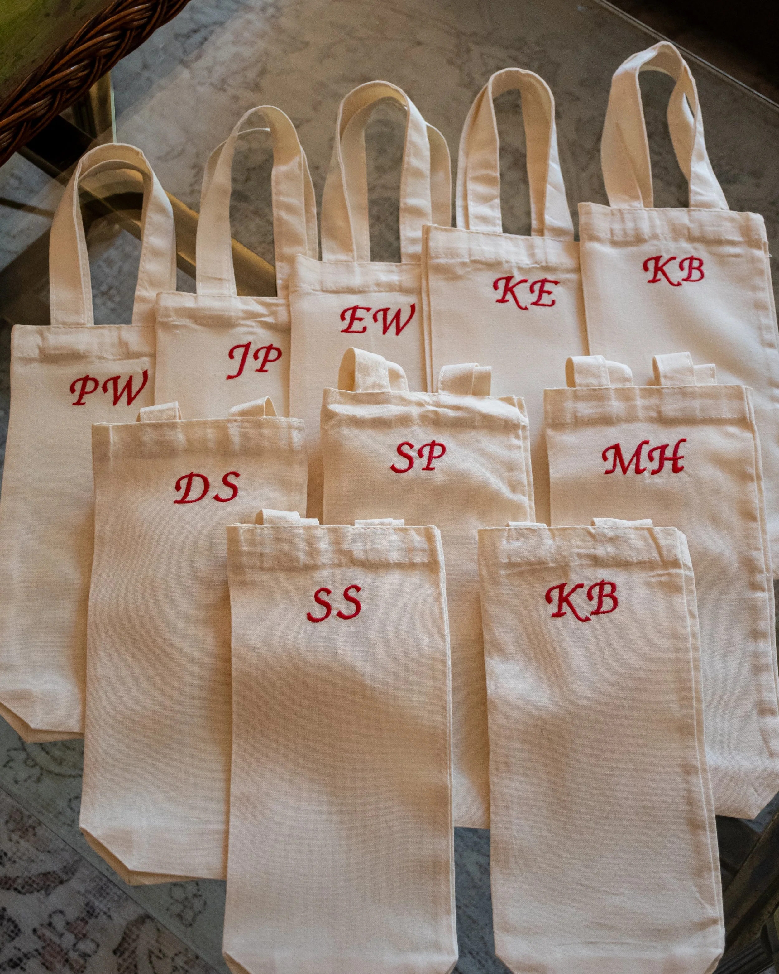 Multiple cream-colored tote bags with red embroidered initials arranged on a glass table, each with two handles.