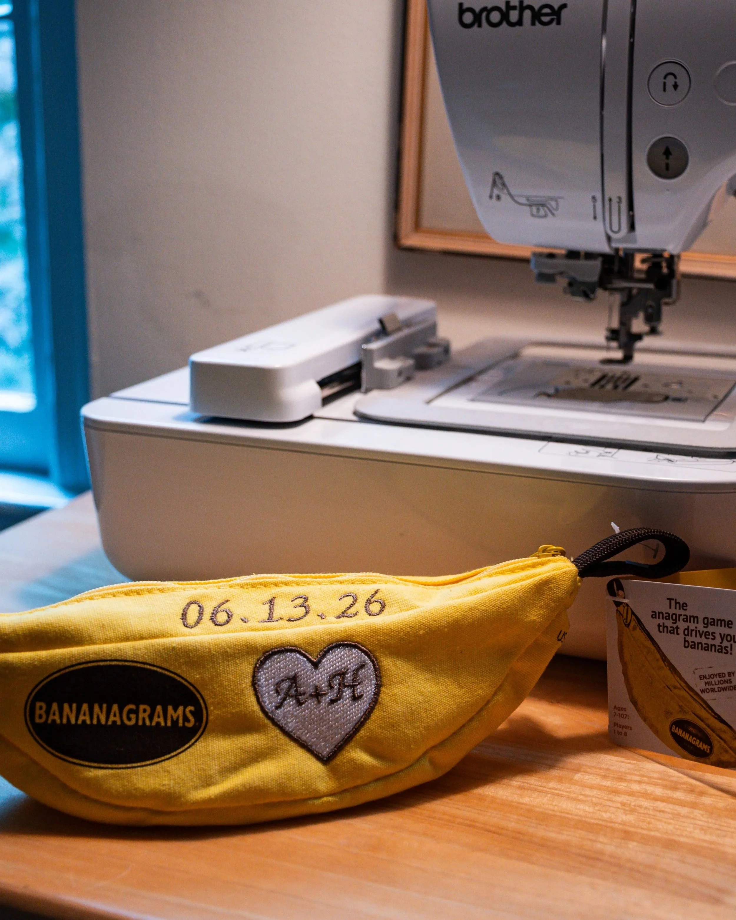A yellow fabric pouch with embroidered text showing the date 06.13.26, sitting on a wooden surface. The pouch has a heart and paw print design and the words "BANANAGRAMS" on it, and is partially covering a pen or marker.