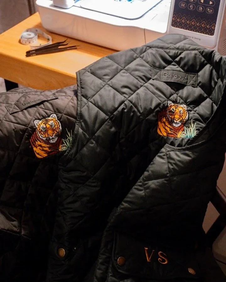 Two black quilted jackets with tiger patches on the chest, hanging on a chair in front of a sewing machine. A wooden table with sewing supplies is in the background.