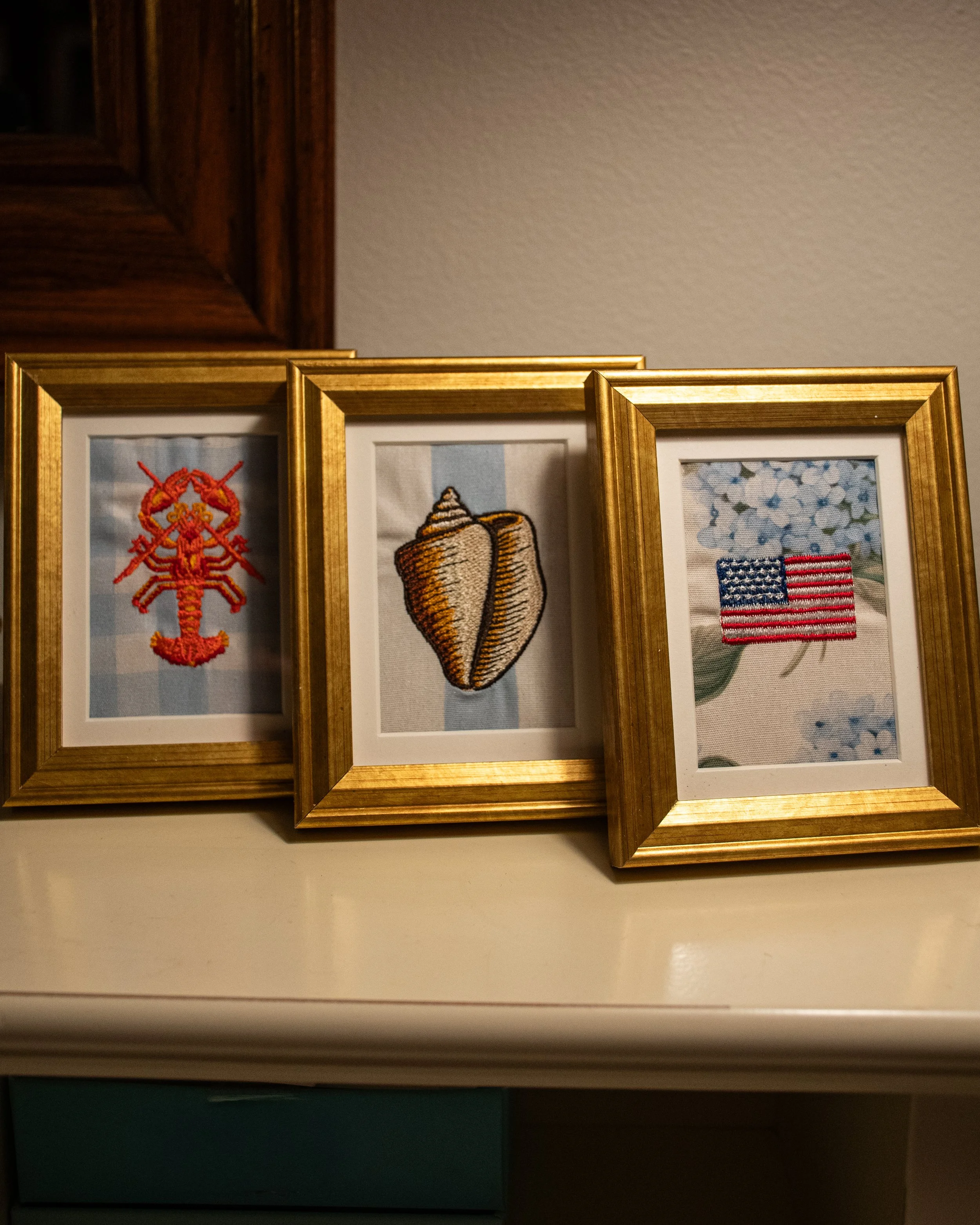 Three framed embroidered patches, including a red lobster, a conch shell, and the American flag, arranged side by side on a white surface against a beige wall.