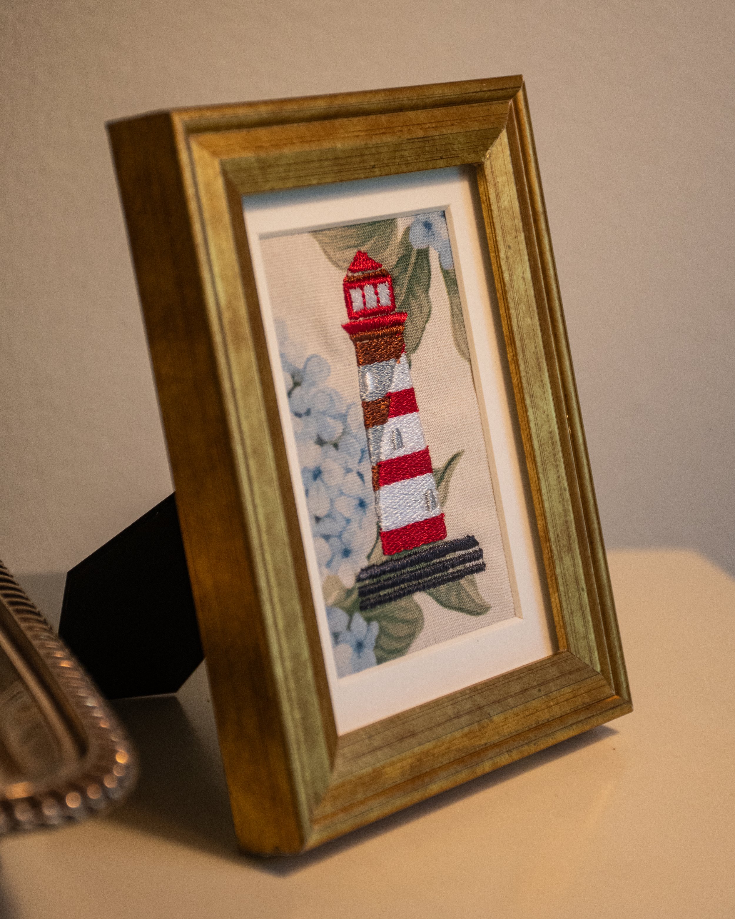 Framed embroidered picture of a lighthouse with red and white stripes, surrounded by blue flowers and green leaves.