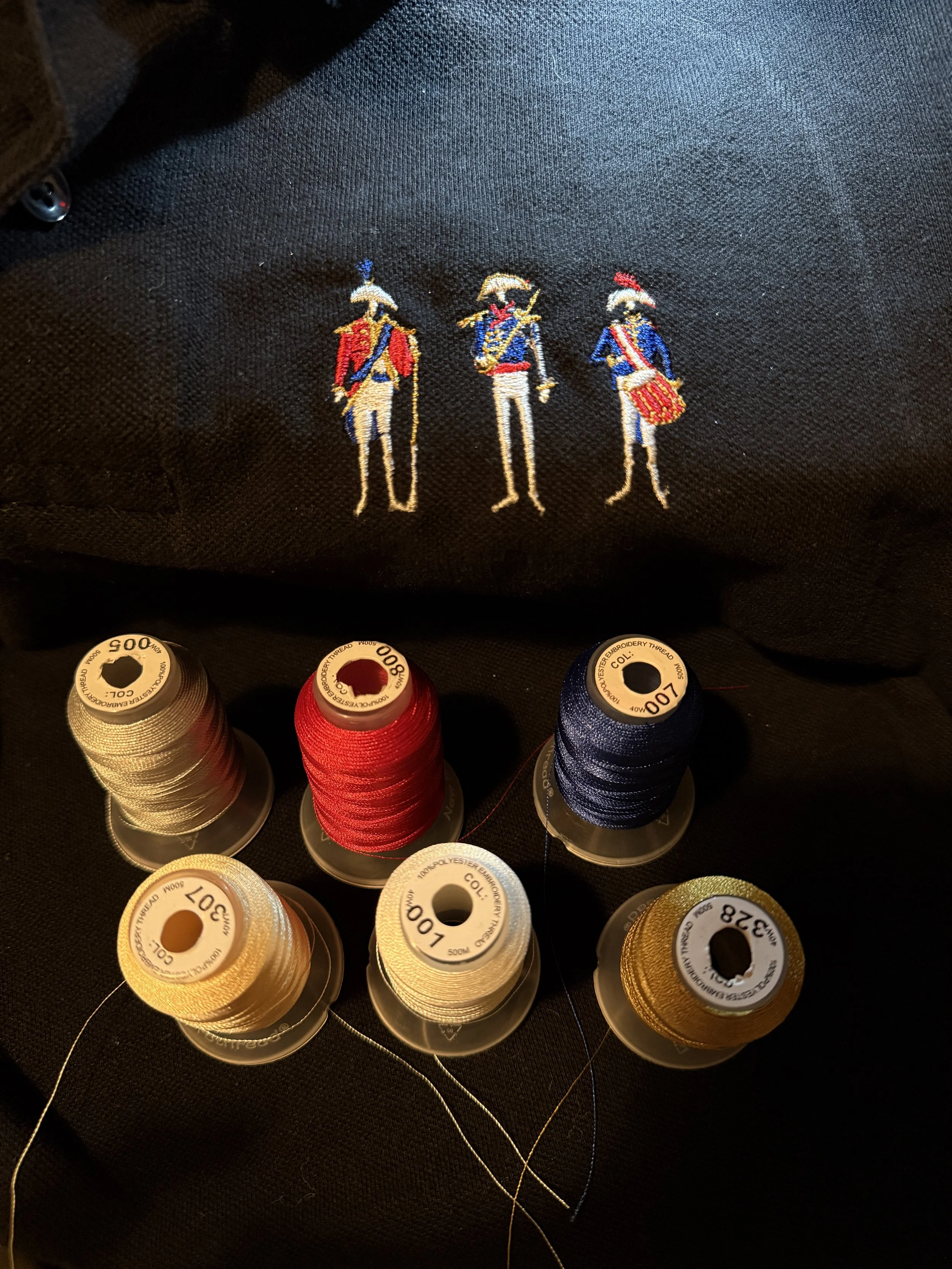 Embroidery with three soldiers in 18th-century military uniforms and six spools of thread in beige, red, navy, and gold colors.