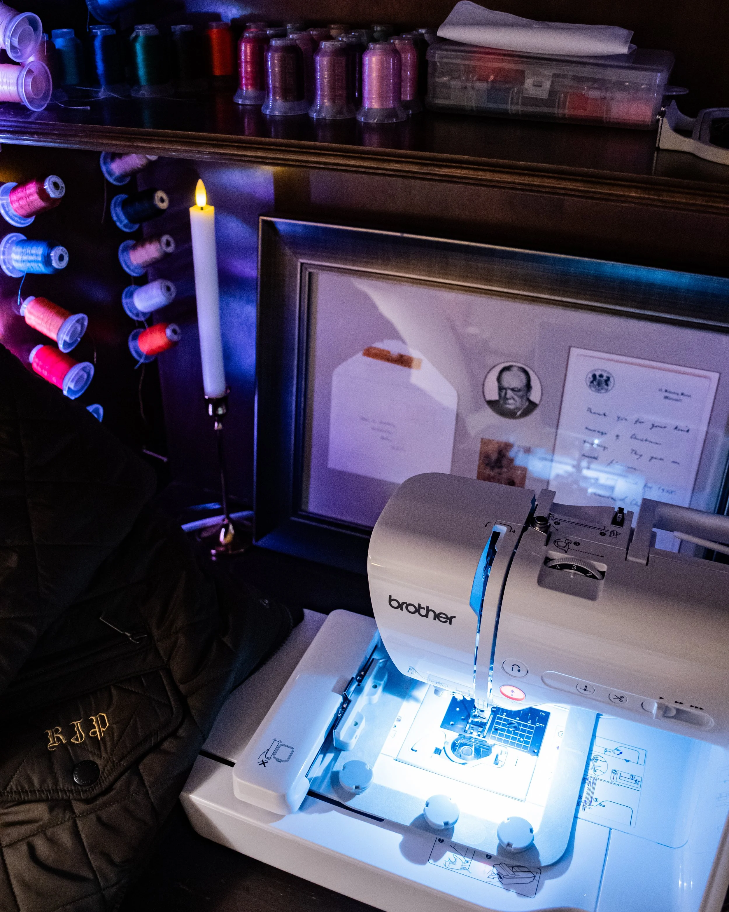 Embroidery machine on a work table with spools of thread and framed certificates or photos in the background.