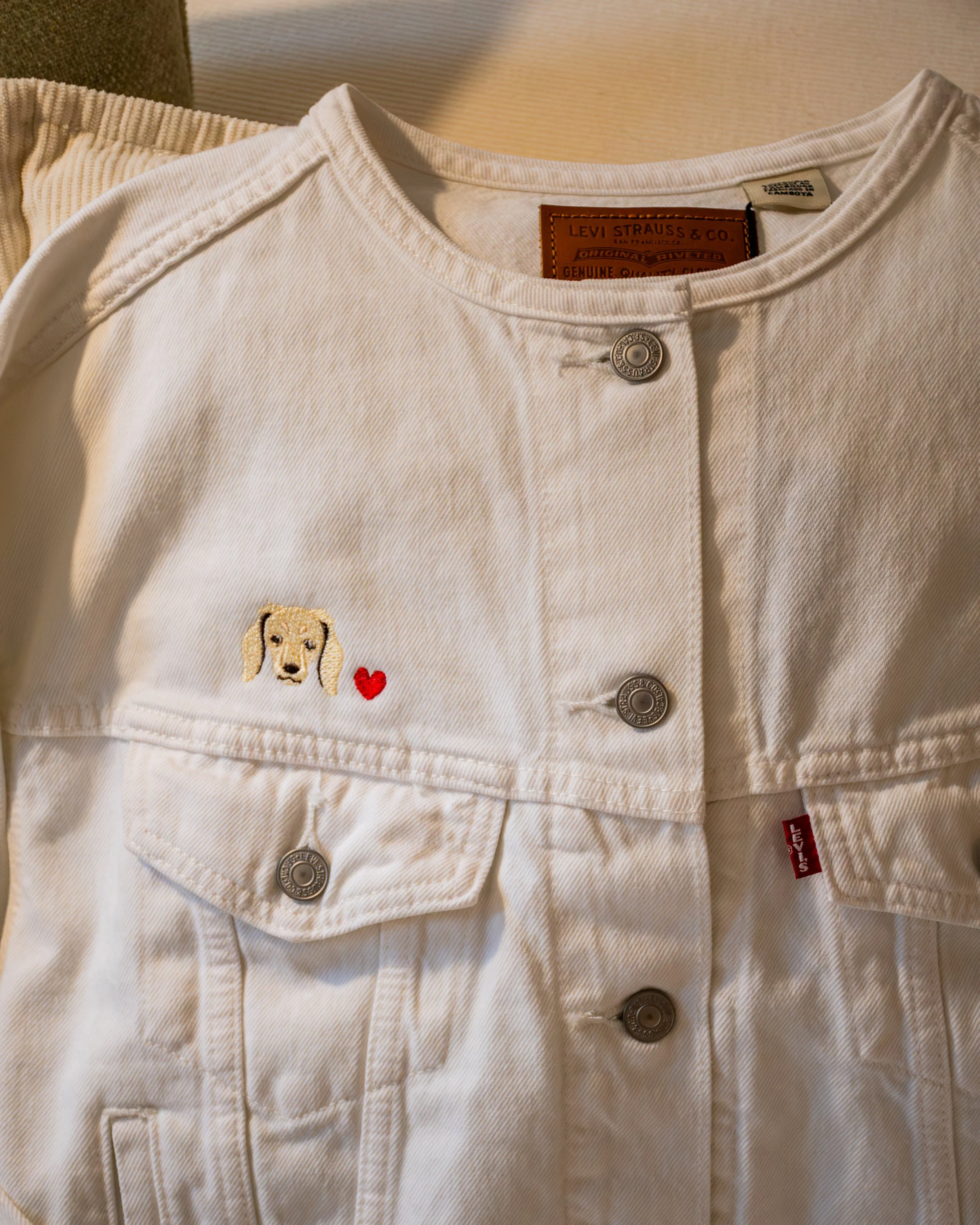 Close-up of a white Levi's denim jacket with metal buttons, embroidered dog patch, and red Levi's tag, laid on a beige fabric surface.