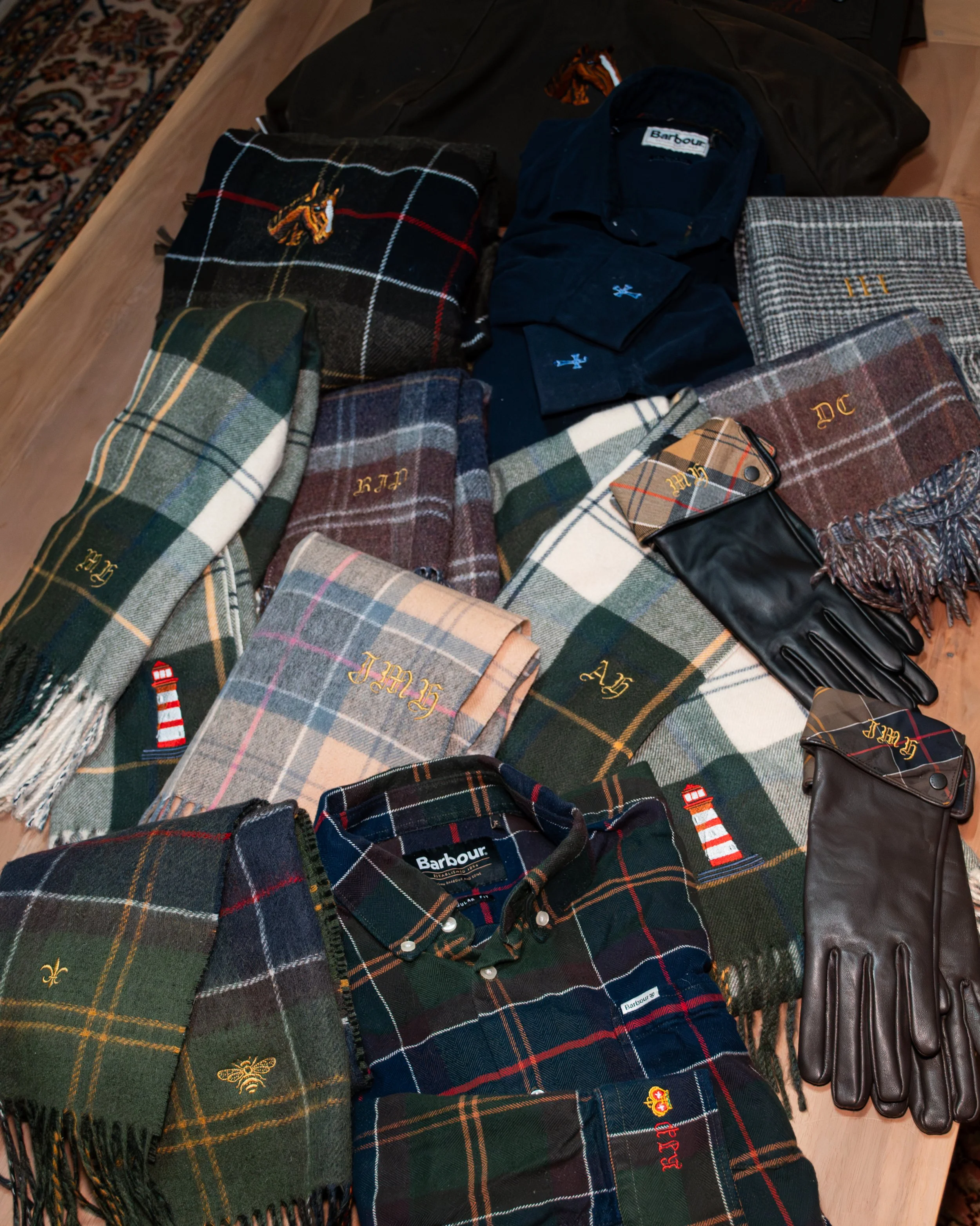A collection of plaid scarves and hats laid out on a table, some with monograms and labels, along with a pair of black leather gloves and a small plaid wallet.