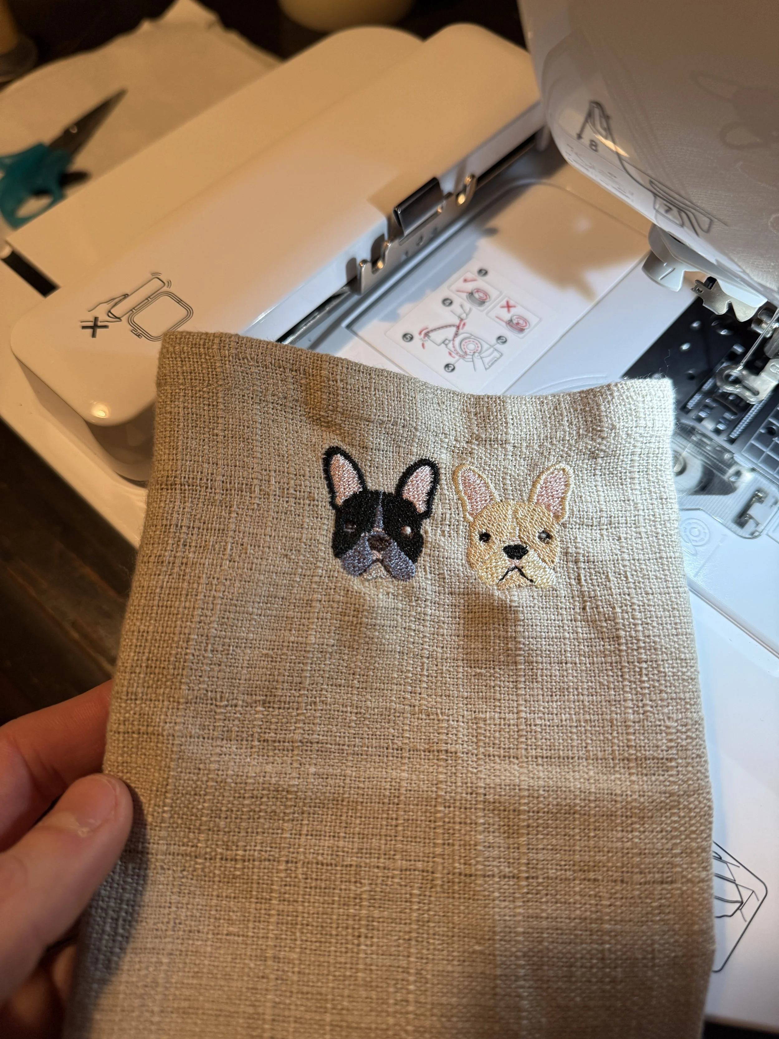 Close-up of a beige fabric with embroidered French Bulldog faces in black and cream, held in front of a sewing machine.