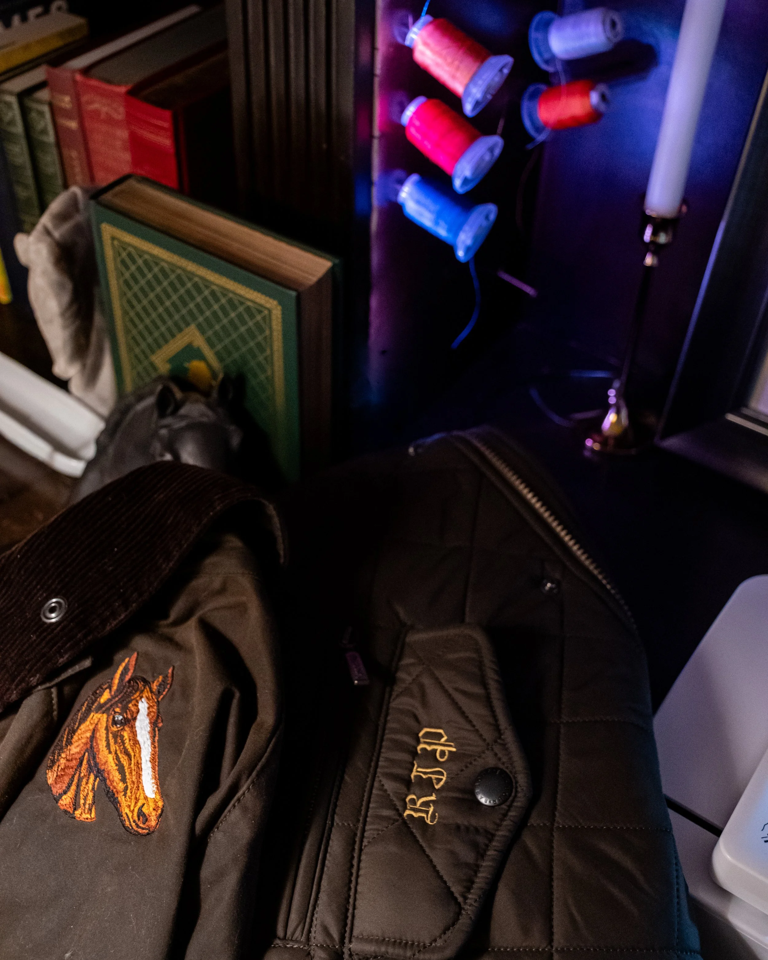 A black quilted jacket with a tiger patch on the sleeve and gold embroidered text that appears to spell 'BE JUNK' or similar. The jacket is open, showing a pair of brown boots, and the person is sitting with their legs crossed. In the background, there is a row of books, some wires, and a wall with colorful spool lights.