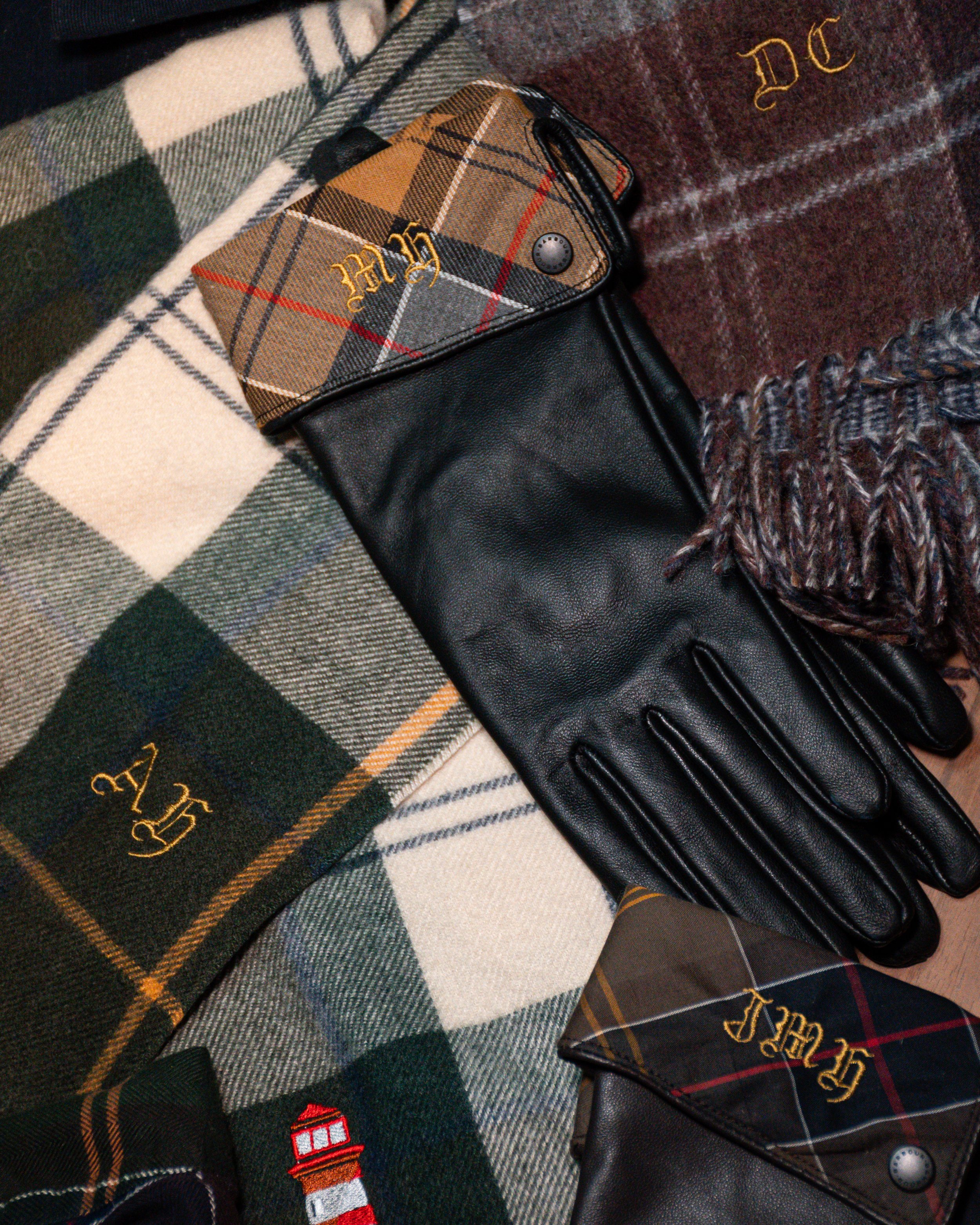 Black leather glove lying on plaid blankets with monograms, one with a Burberry check pattern and embroidered initials.
