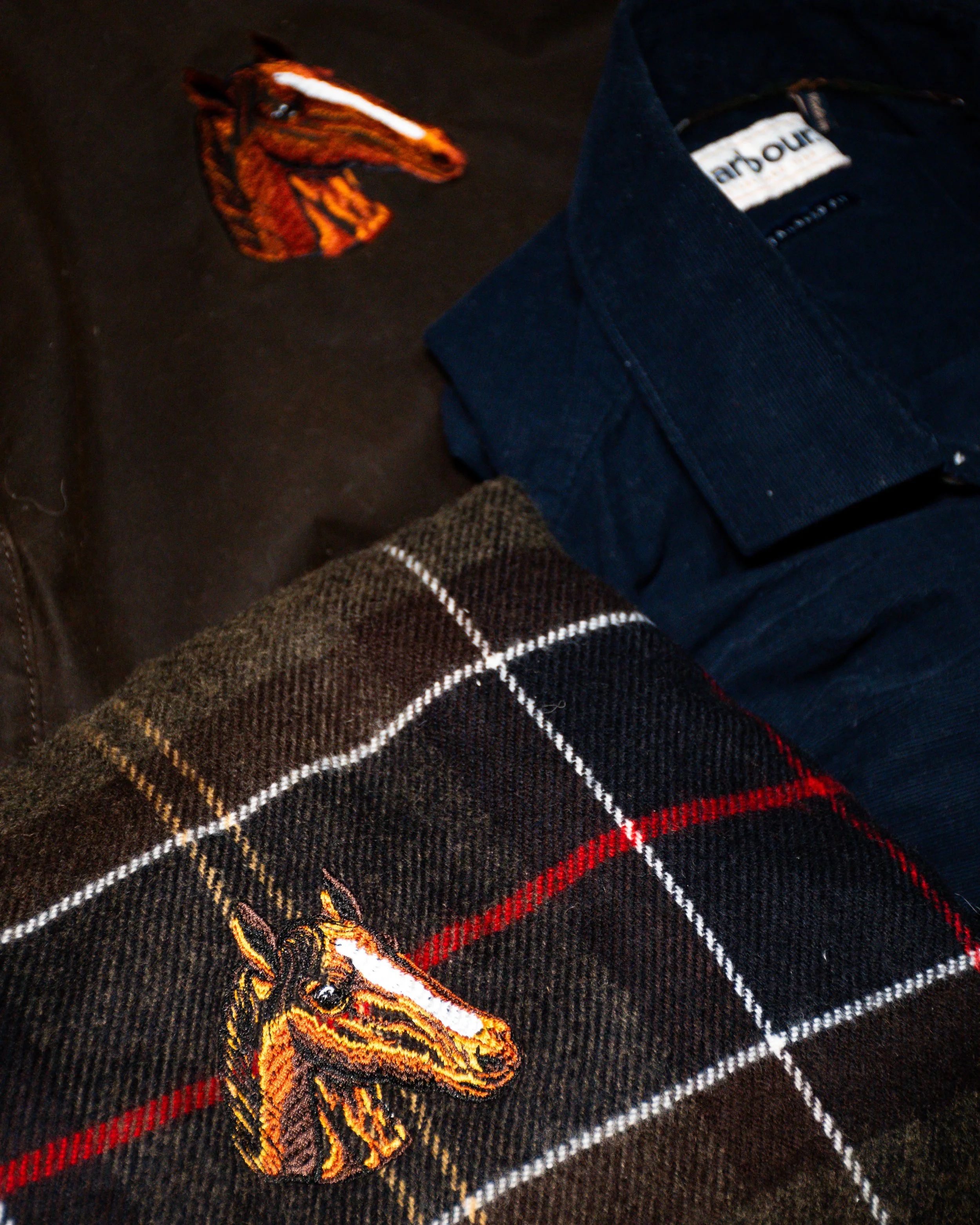 Close-up of a fabric with an embroidered horse head patch, a folded plaid scarf, and a dark blue denim shirt.