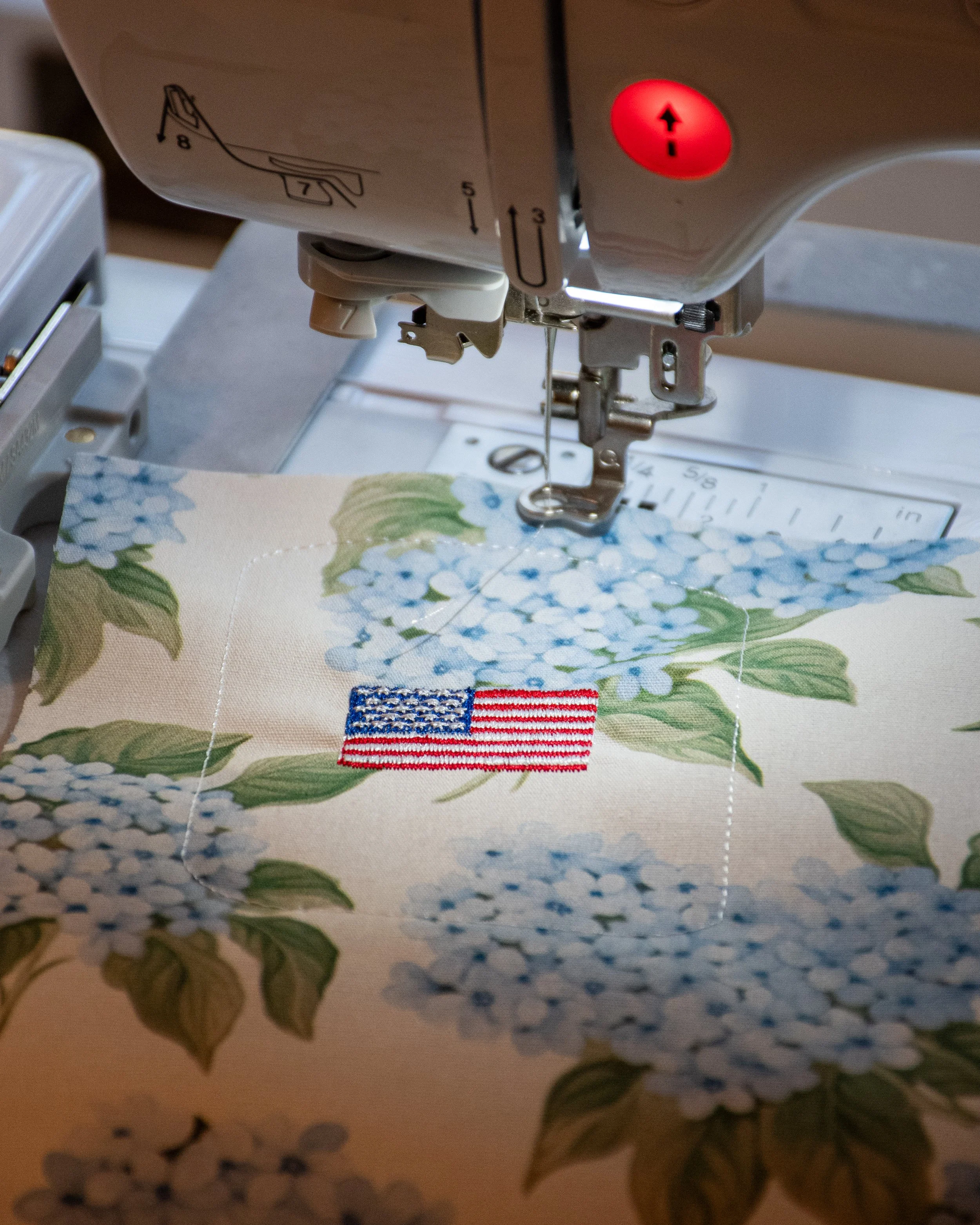 A sewing machine stitching an American flag patch onto fabric with a floral print.