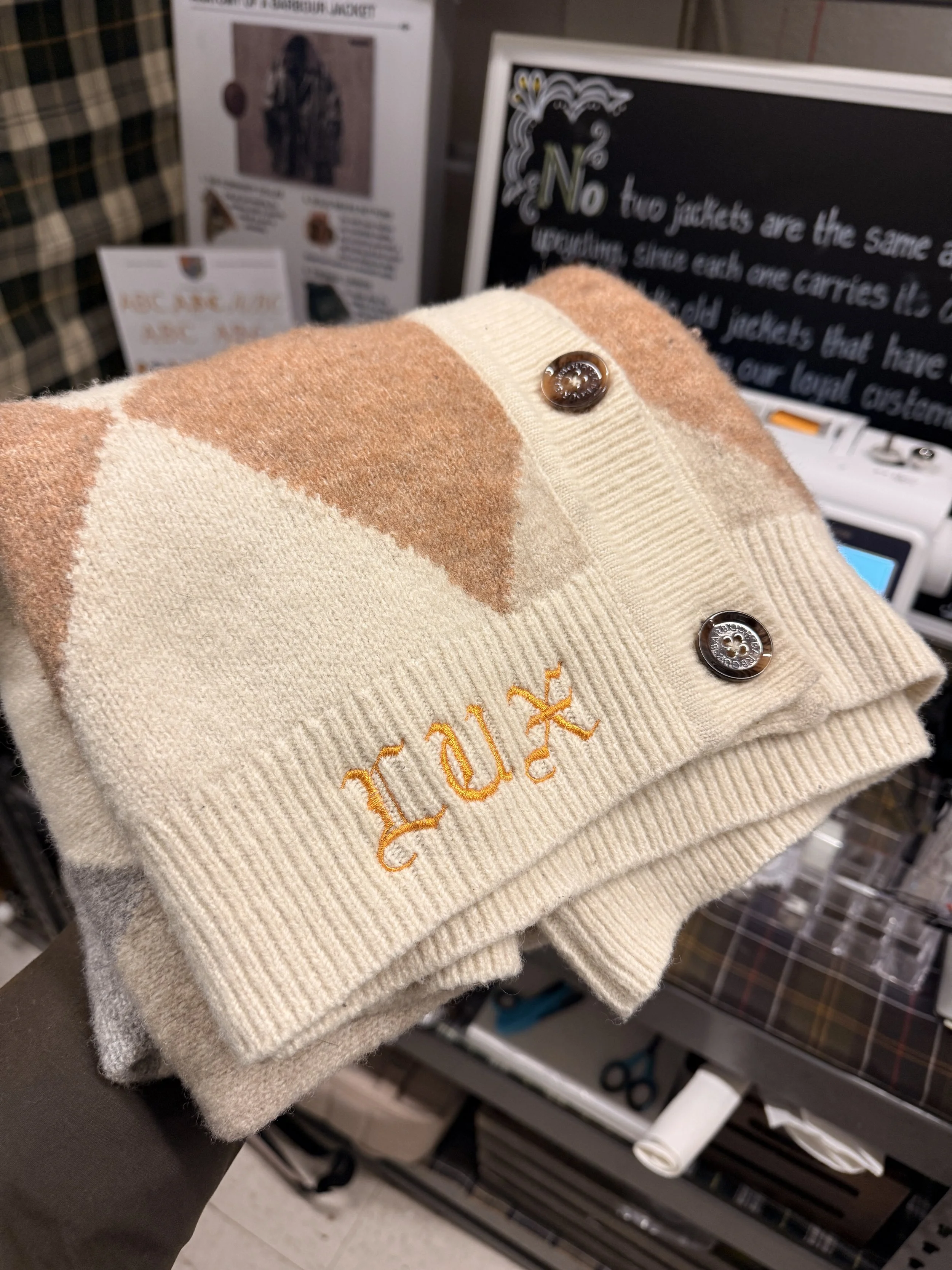 Close-up of a folded cream-colored sweater with orange embroidery and two brown buttons, on display at a store. In the background, a blackboard with white text and other store items are visible.