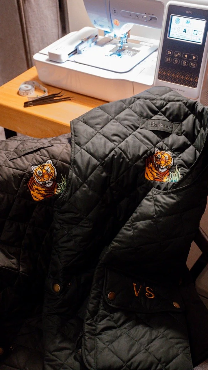 Two black quilted jackets with embroidered tiger patches on the chest, placed on a leather chair in front of a sewing machine on a wooden table.