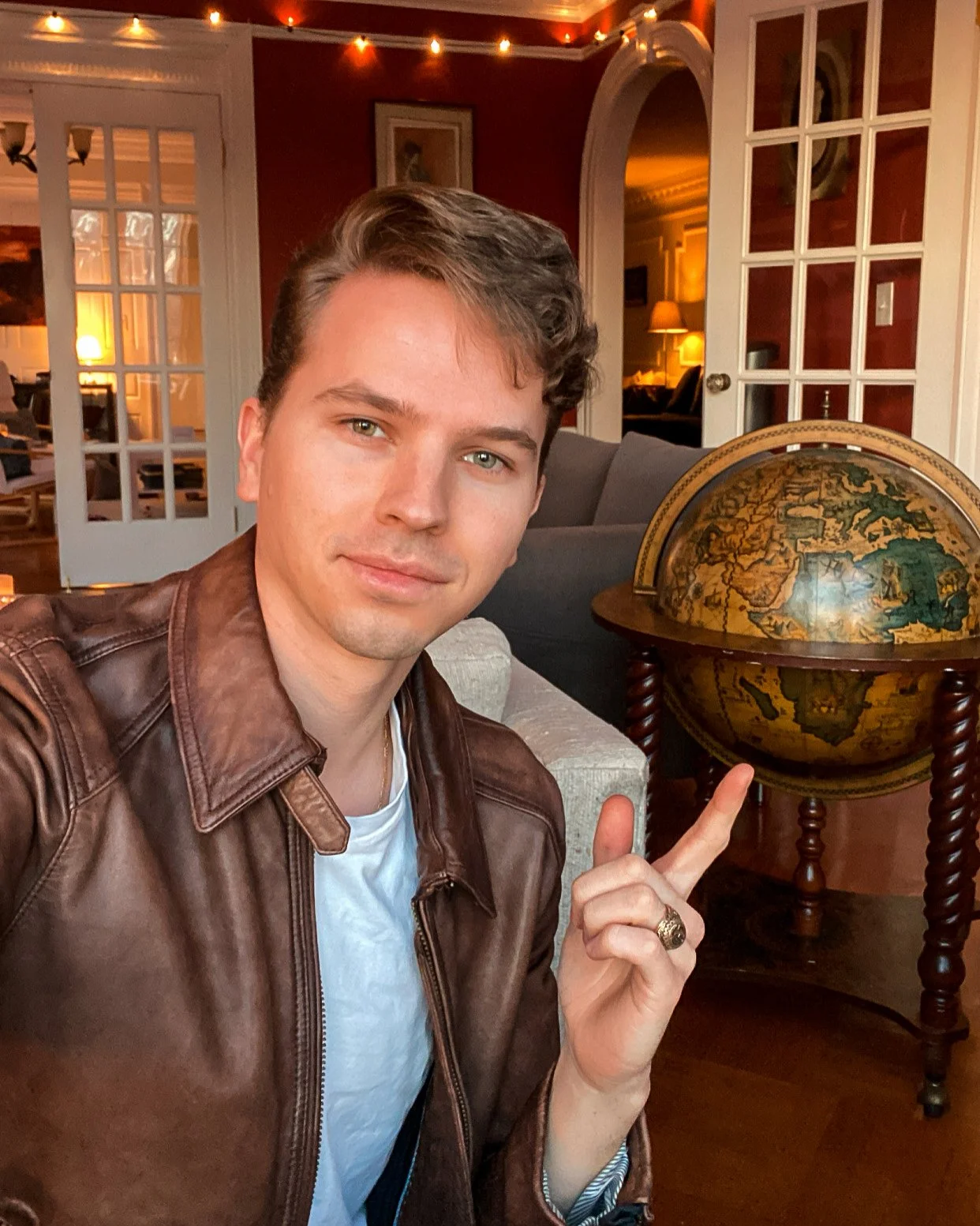 A young man with brown hair wearing a brown leather jacket and white shirt points to a vintage globe on a wooden stand in a cozy living room with warm lighting and string lights.