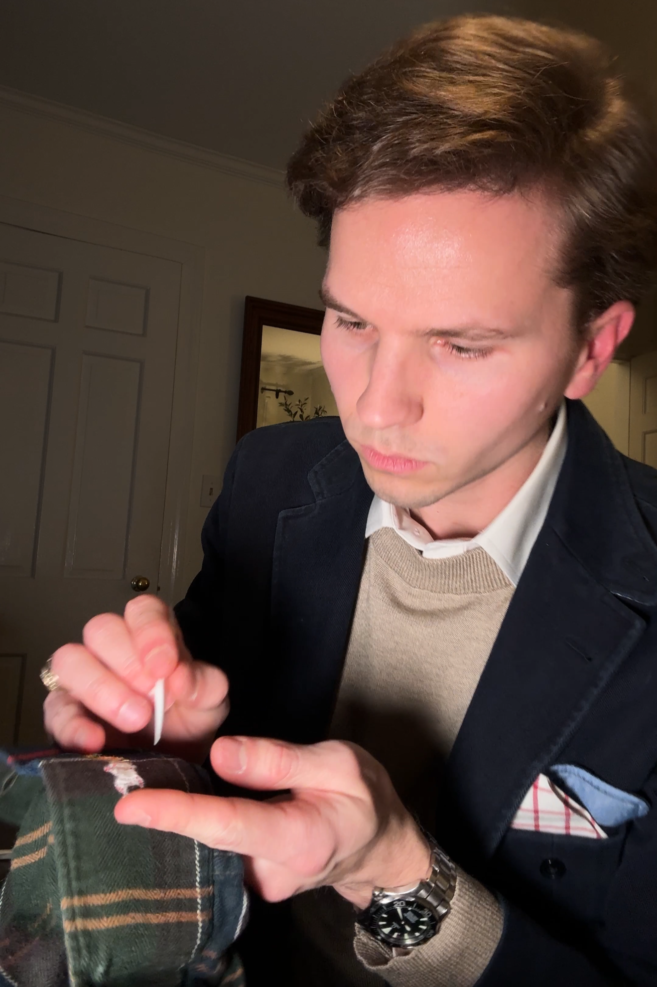 A young man with brown hair, dressed in a dark jacket and a beige sweater, is applying a white tattoo to the person's hand with a tattoo needle.