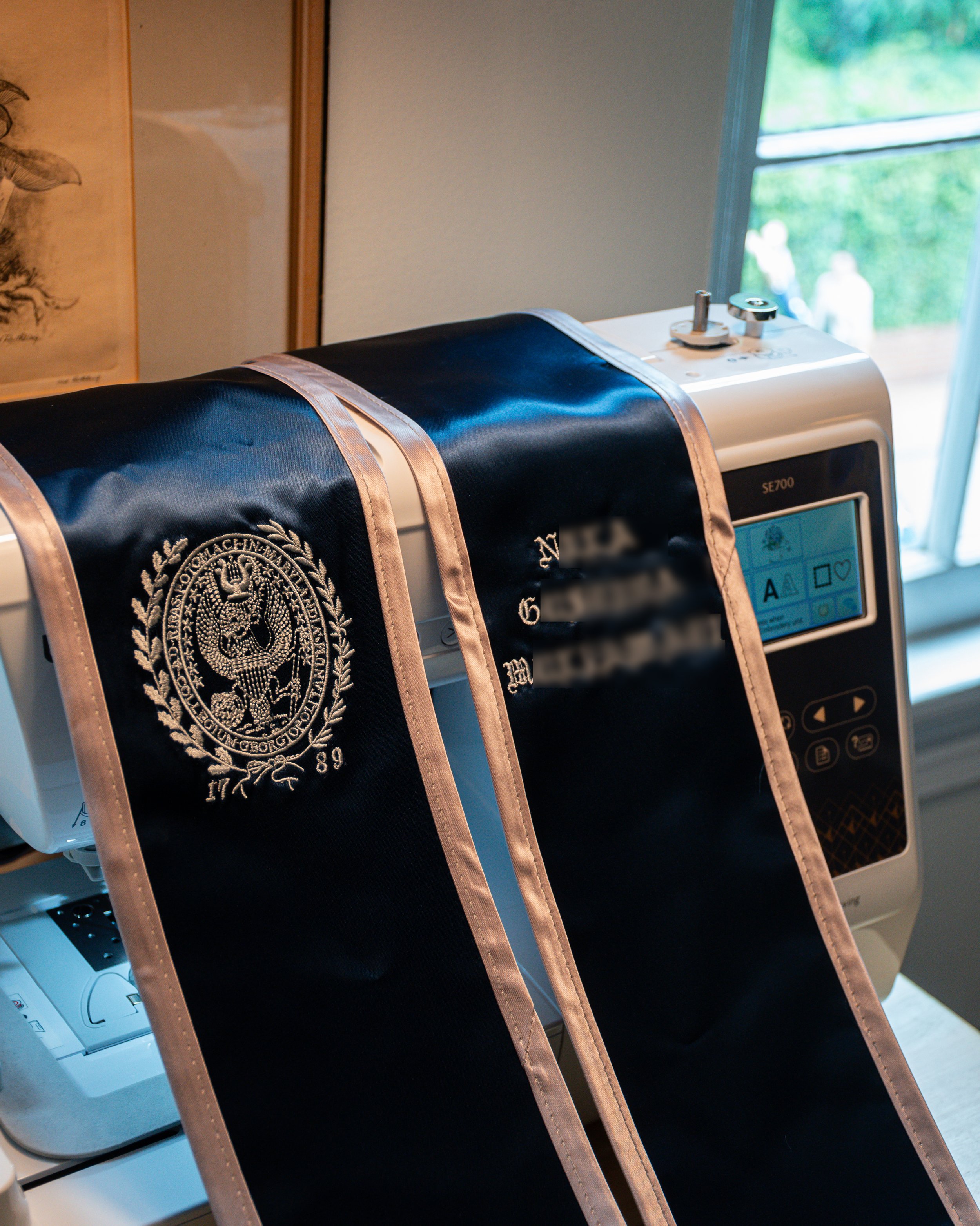 Close-up of an embroidery machine with an embroidered graduation sash draped over it, showing a detailed emblem and text, in front of a window with blurred outdoor scenery.