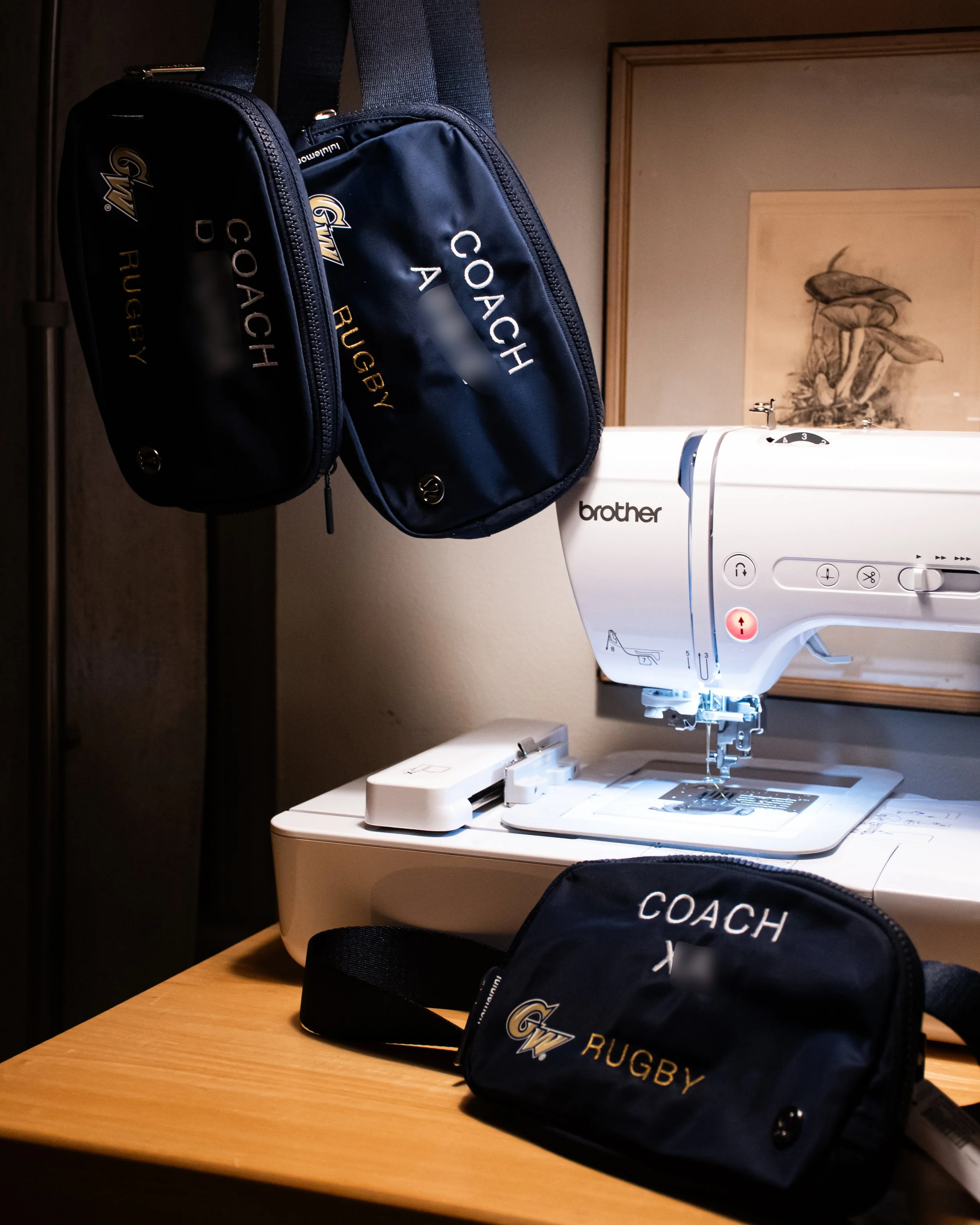 A sewing machine on a wooden table with a Lululemon hip bag in front and another embroidered rugby bag hanging on a hook, in a room with wooden molding and framed artwork on the wall.