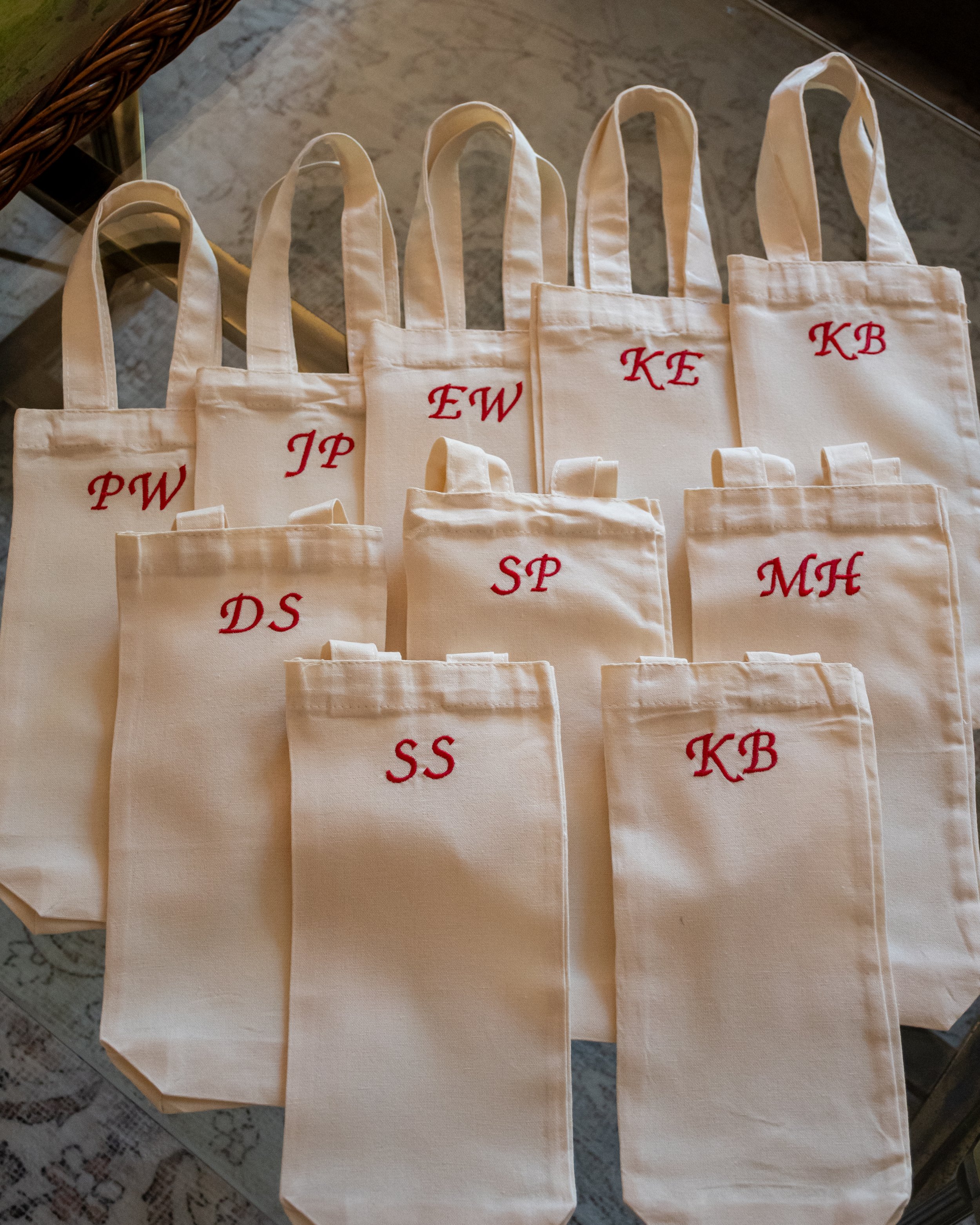 Multiple white canvas tote bags with red embroidered initials, arranged on a table. The initials are PW, JP, EW, KE, KB, DS, SP, and MH.