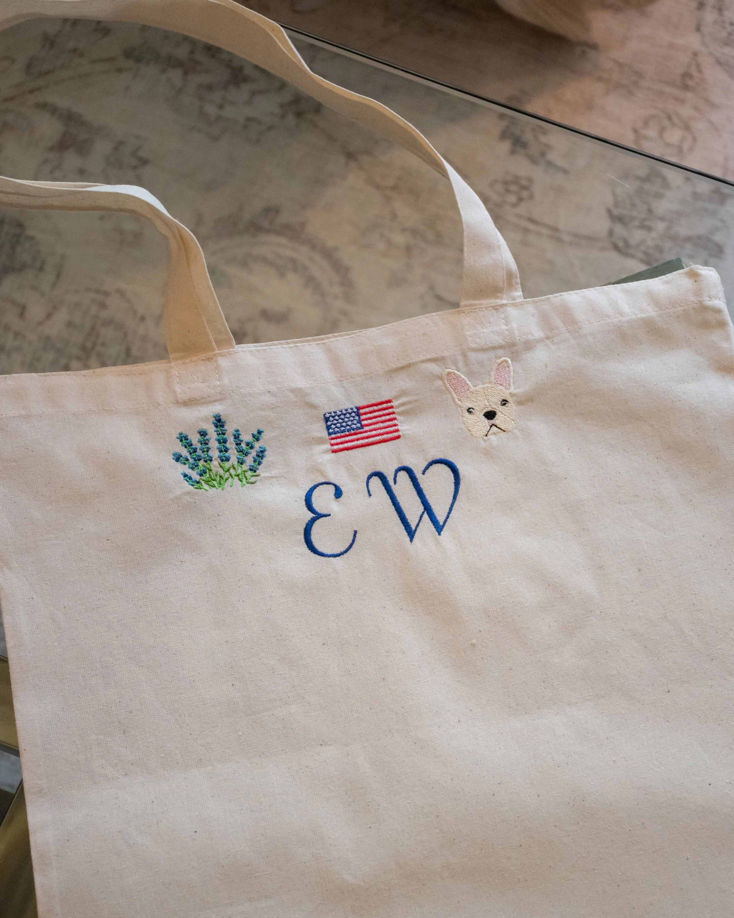 Close-up of a beige tote bag with embroidered trees, an American flag, and a French Bulldog face, hanging on a glass-topped wooden surface.