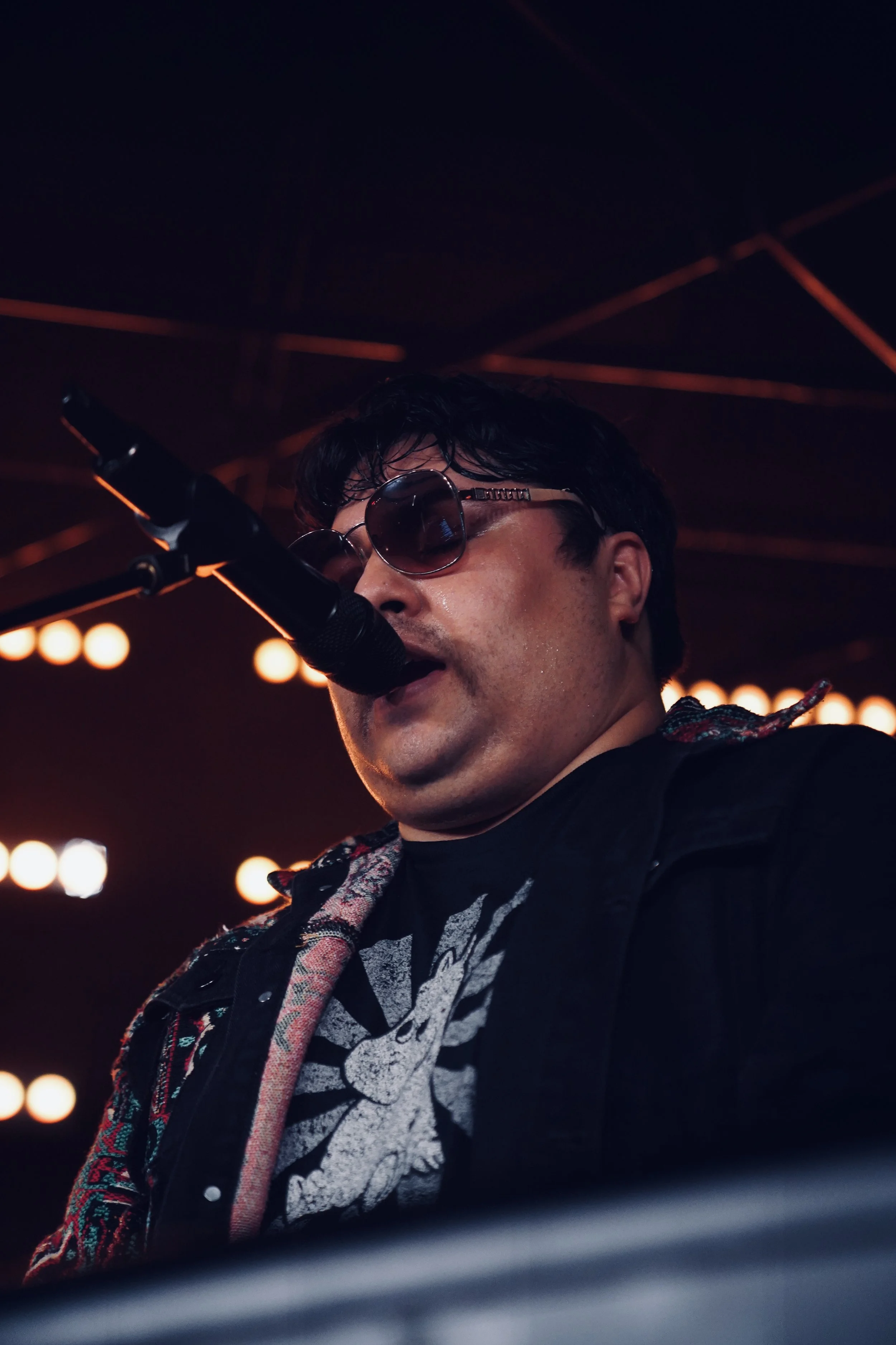 A male singer wearing sunglasses performs with a microphone on a stage lit with warm lighting.