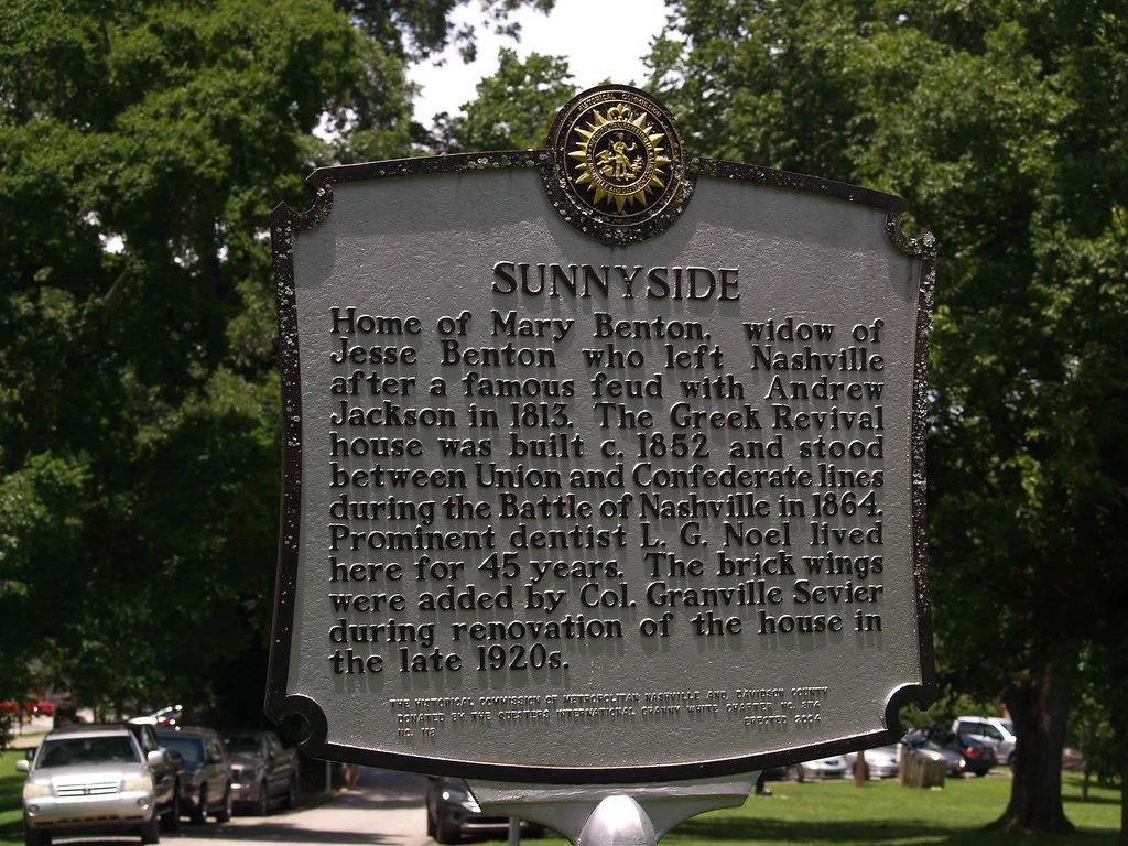Historical plaque for Sunny Side, the house of Mary Benton, widow of Jesse Benton. Features a gold emblem at the top, with green trees and parked cars in the background.