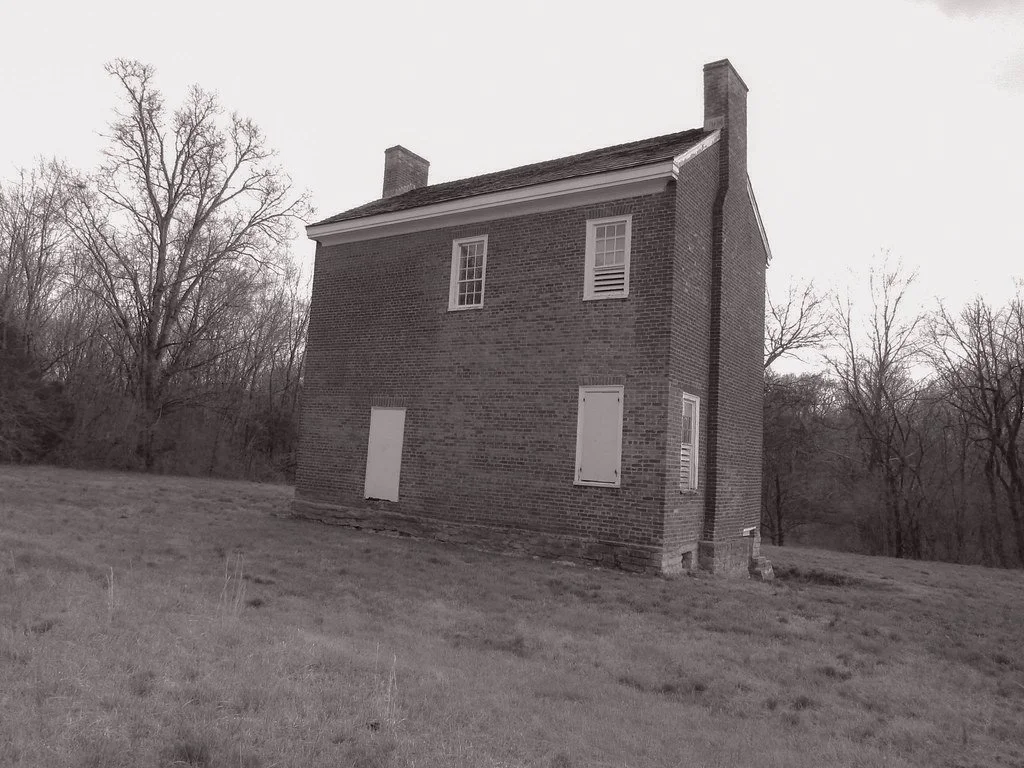 A two-story brick house with boarded-up windows on a grassy field with leafless trees in the background.