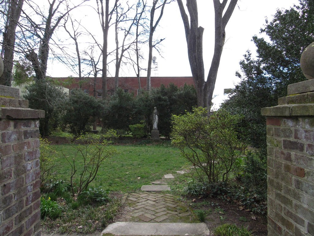 A garden with brick pillars on each side, leading to a grassy area with bushes and trees, and a statue of a person at the center in the background.