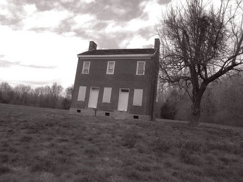An old, abandoned brick house with boarded-up windows and a leafless tree in front, on a grassy field with a cloudy sky.