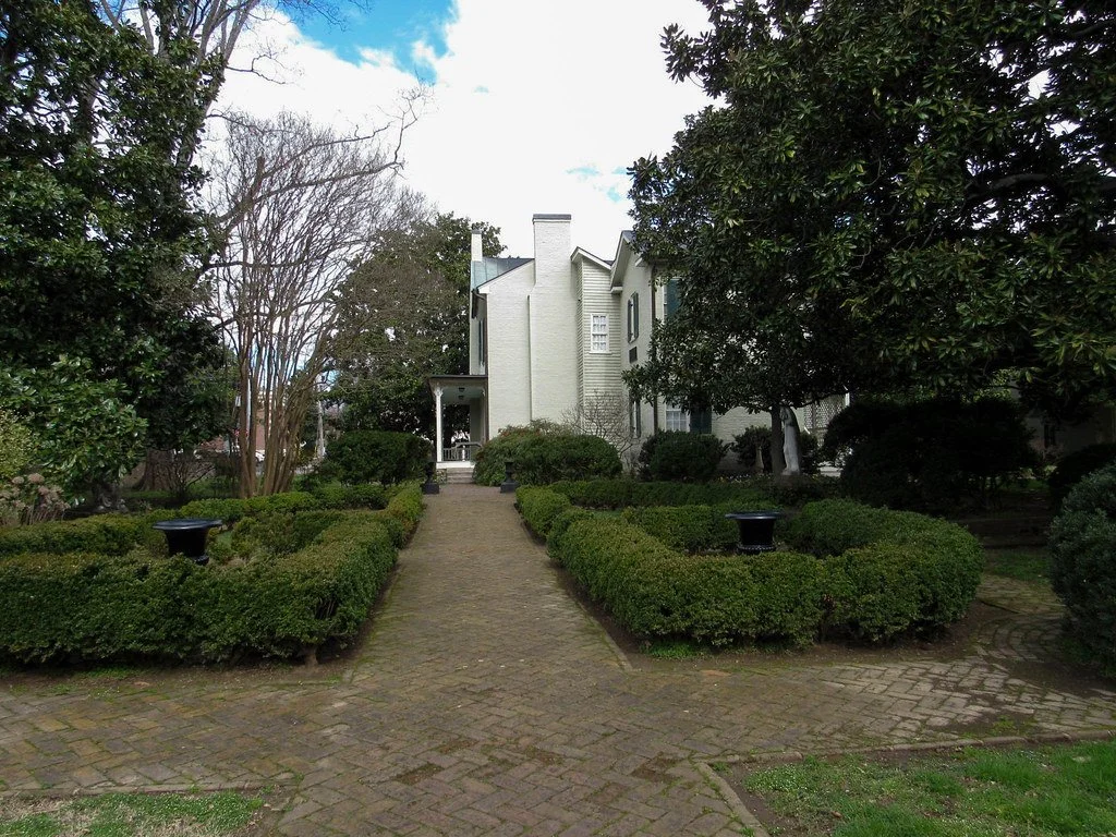 A garden with a brick pathway leading to a white house, surrounded by bushes and trees.