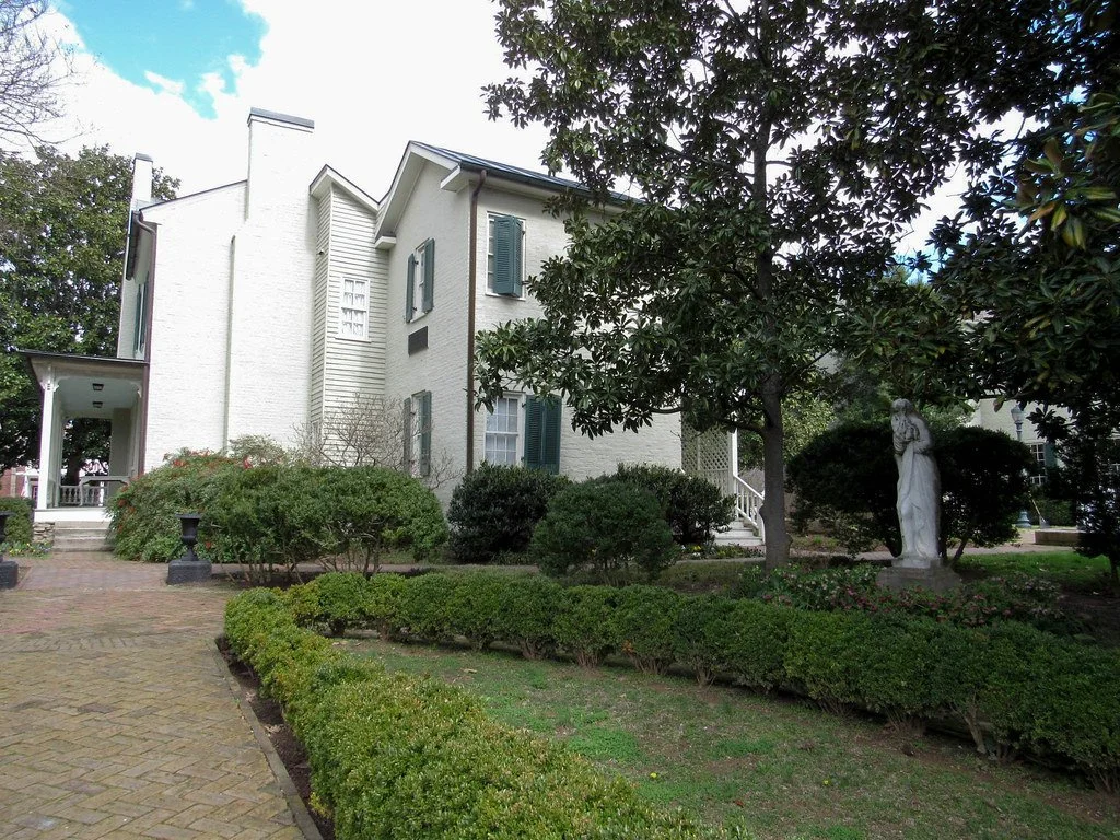A white, multi-story house with green shutters, surrounded by a well-maintained garden with bushes, trees, and a statue of a woman.