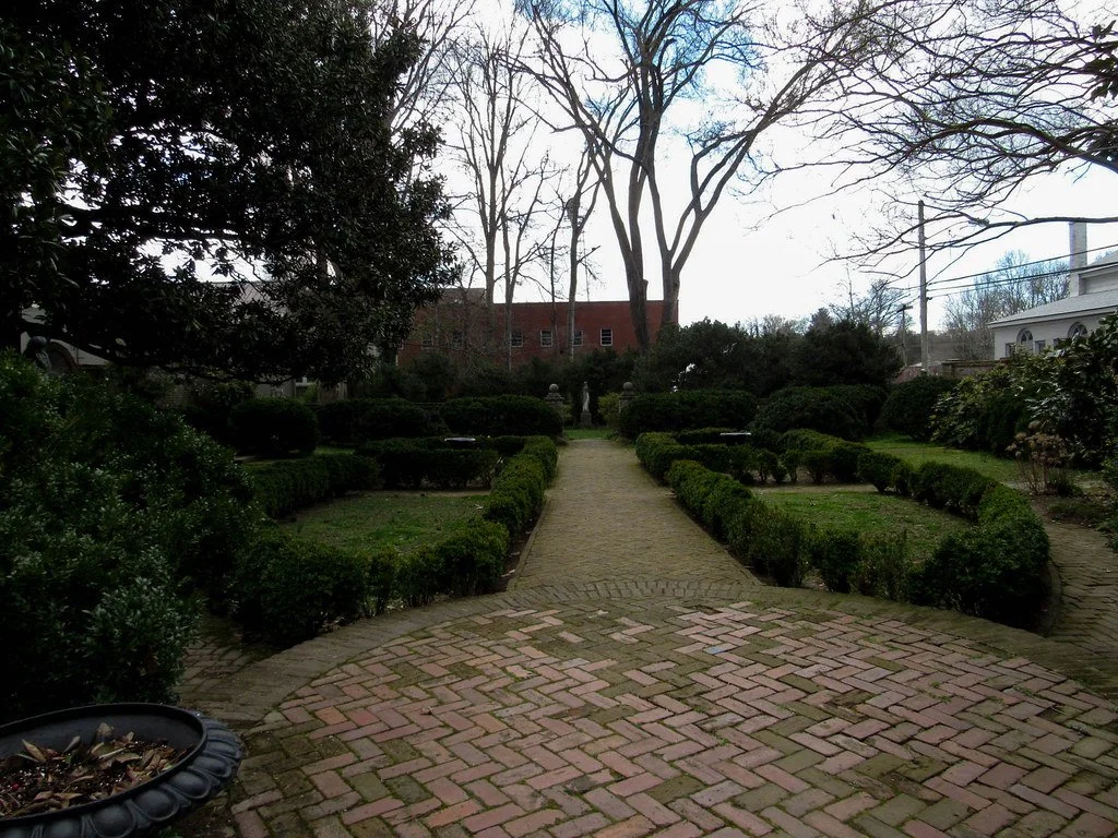 A brick pathway leading through a garden with trimmed bushes on either side, trees in the background, and a cloudy sky overhead.