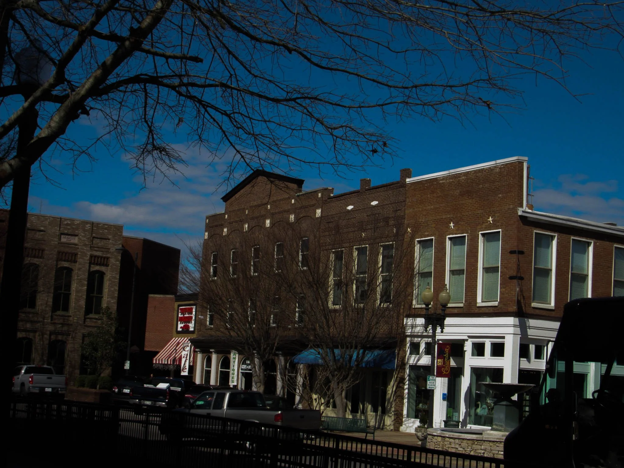 Downtown street scene with brick buildings, parked cars, and leafless trees under a blue sky.