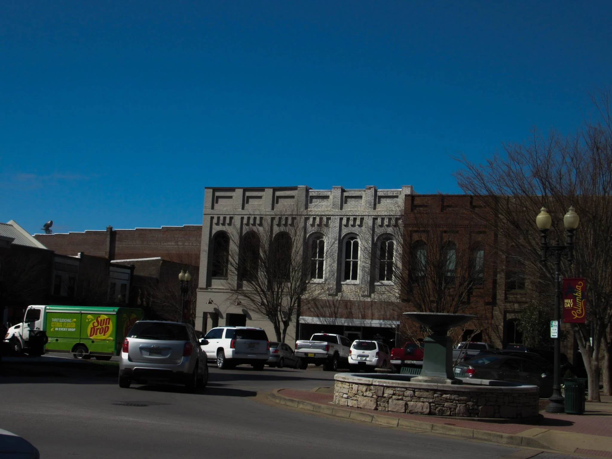 Downtown street scene with parked cars, a water fountain, brick buildings, leafless trees, a banner, and a bright blue sky.