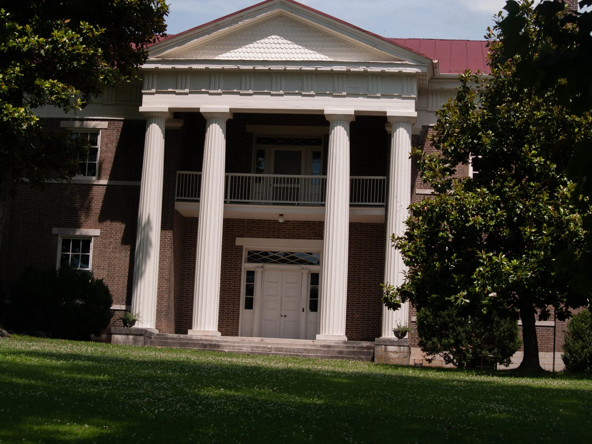 Front view of a large historic brick house with white columns, a balcony, multiple windows, and a well-maintained lawn with trees.