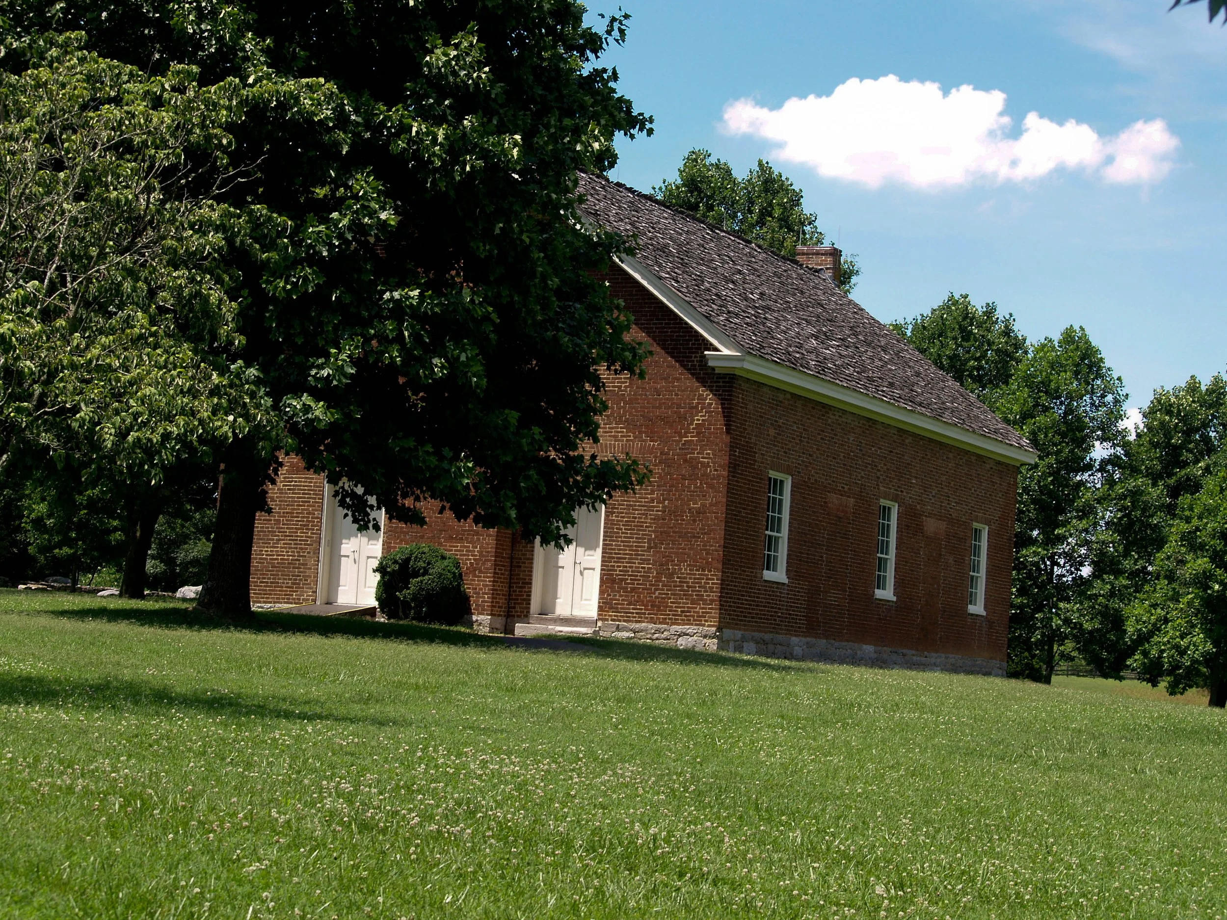 A small brick building with a gabled roof, surrounded by green grass and trees, against a blue sky with a few white clouds.