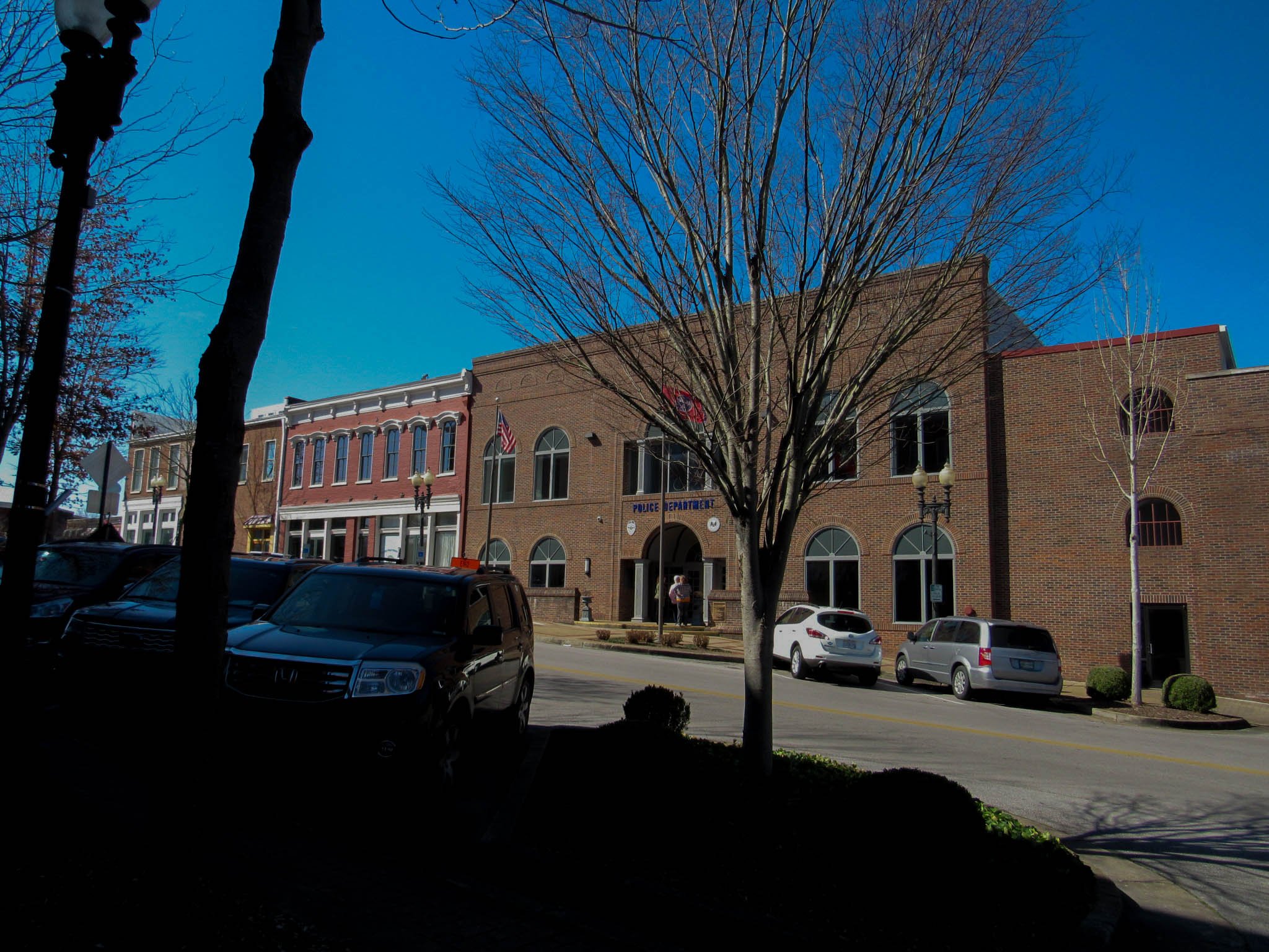 Street view of a downtown area with brick buildings, parked cars, leafless trees, and a building marked as police department under a clear blue sky.