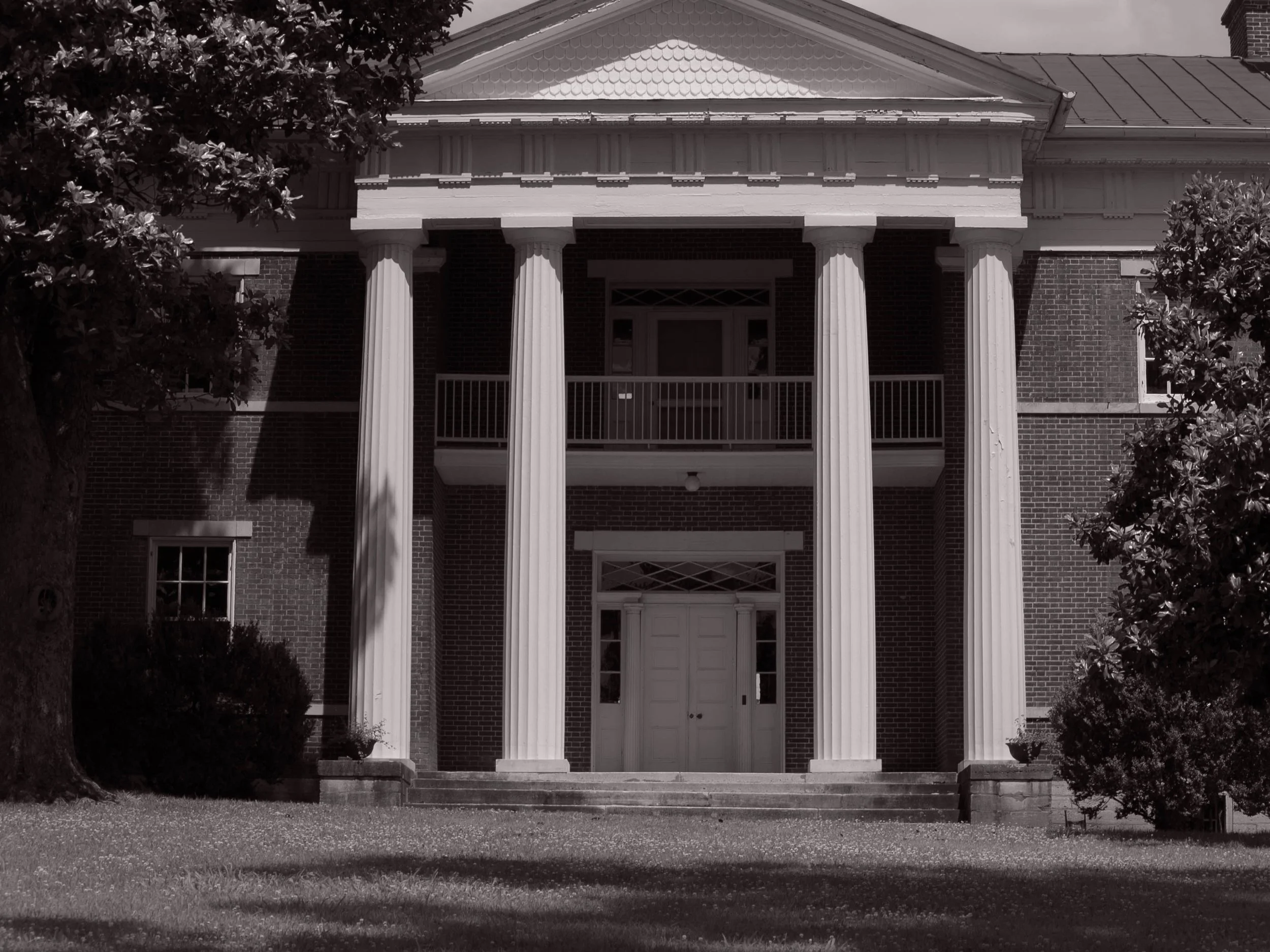Black and white photo of a large, historic brick house with tall white columns, a front porch with stairs, and trees on either side.